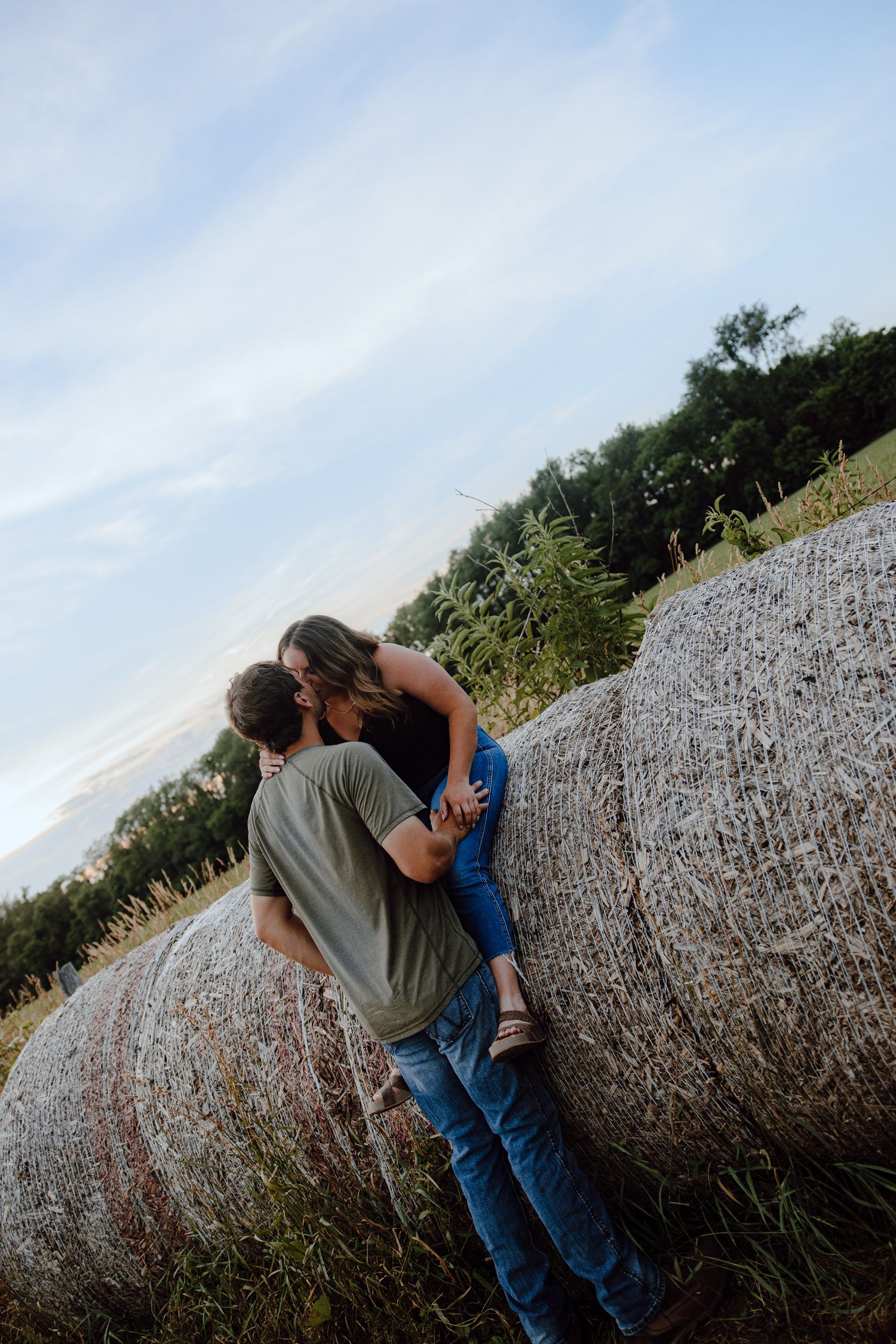 A couple shares a romantic moment against a large round hay bale in a rural field.
