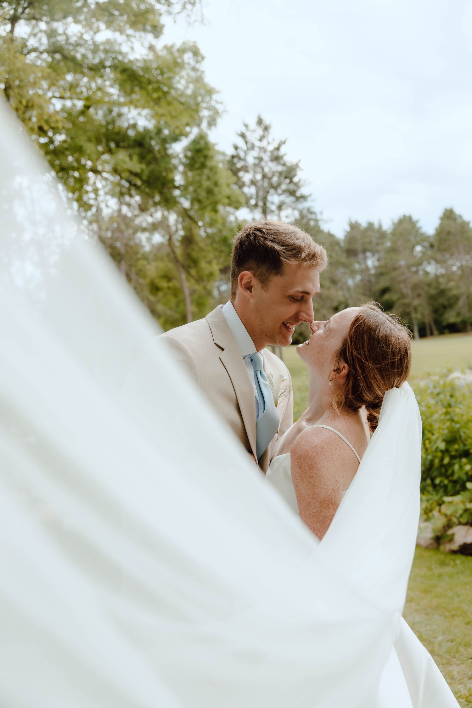 Bride and groom sharing a romantic moment outdoors, framed by a flowing white veil with lush green trees behind them.