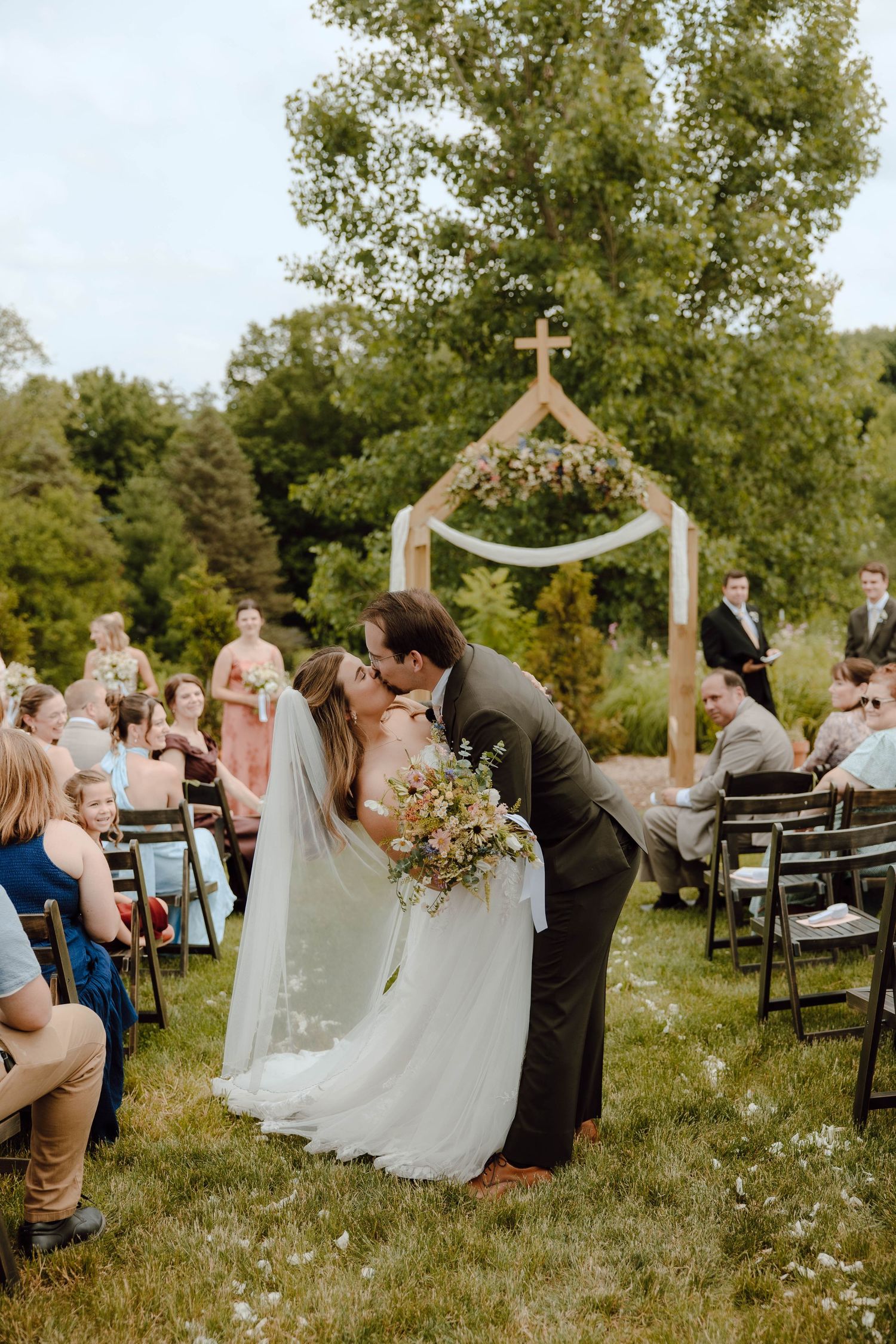 Bride and groom share first kiss at outdoor wedding ceremony with wooden cross arch and seated guests on green lawn.