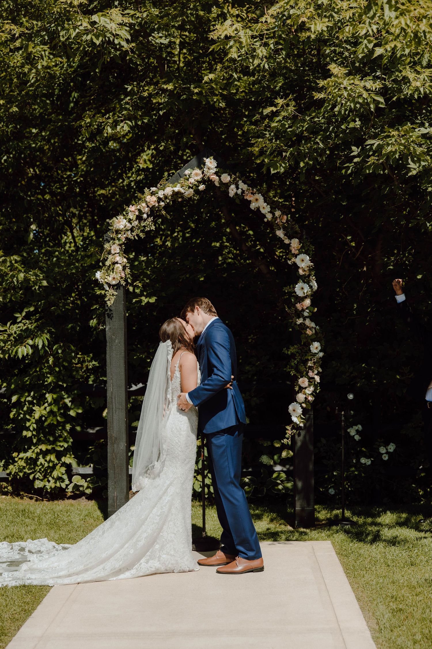 Bride and groom share first kiss under floral arch at outdoor wedding ceremony surrounded by lush green trees.