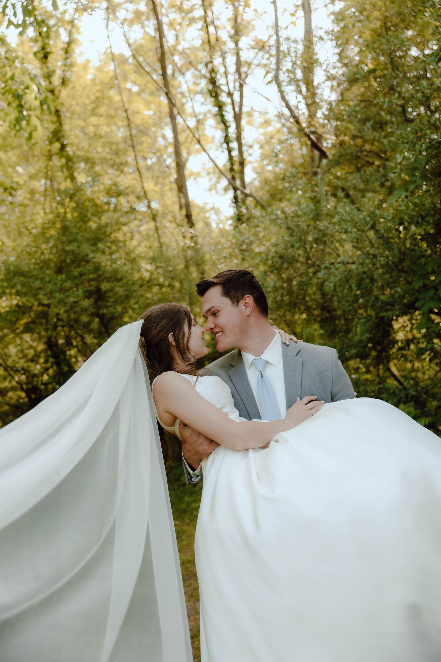 Bride and groom share an intimate moment as her flowing veil billows around them in a sunlit garden setting.