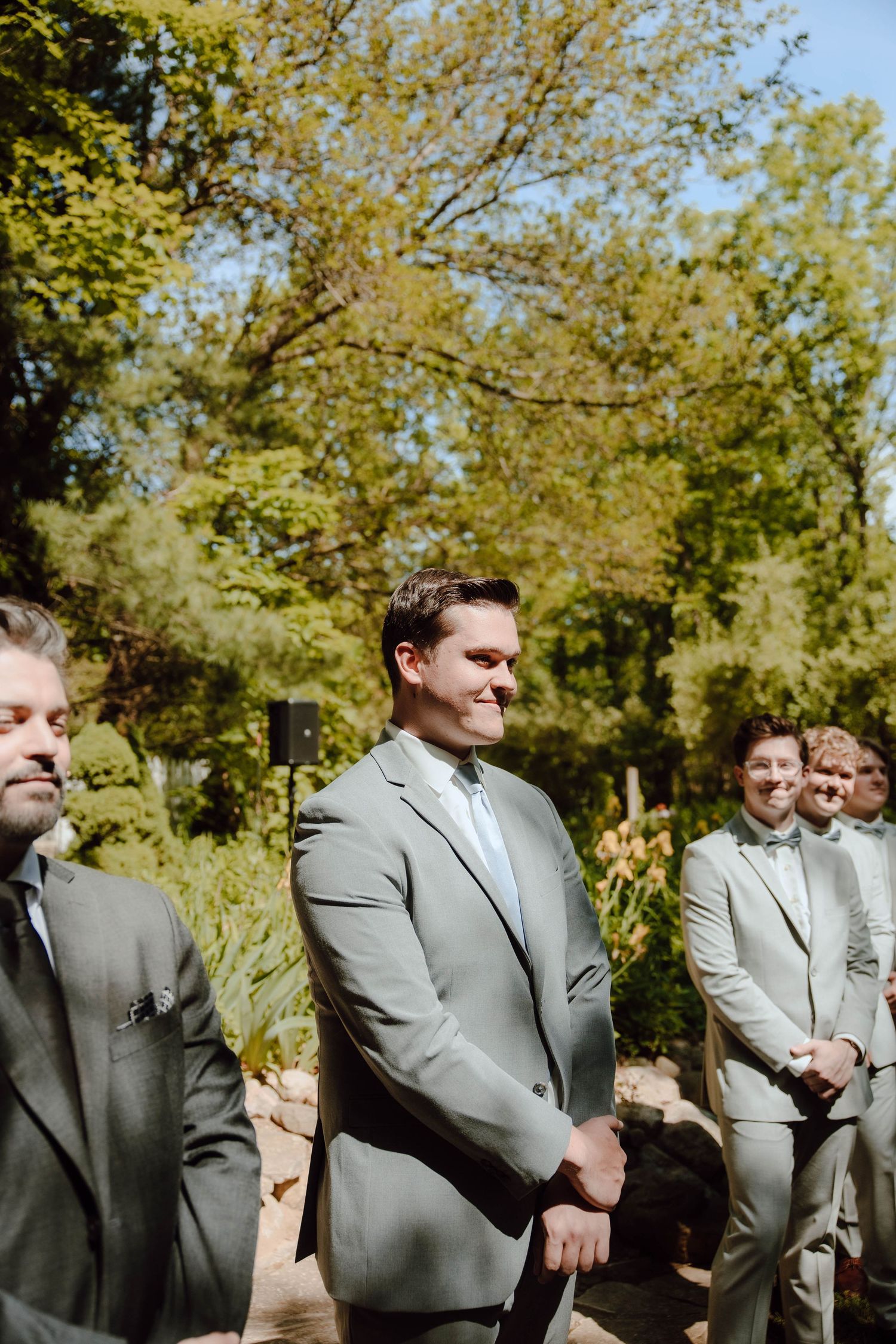 Groomsmen in light gray suits smile during an outdoor wedding ceremony surrounded by lush green trees.