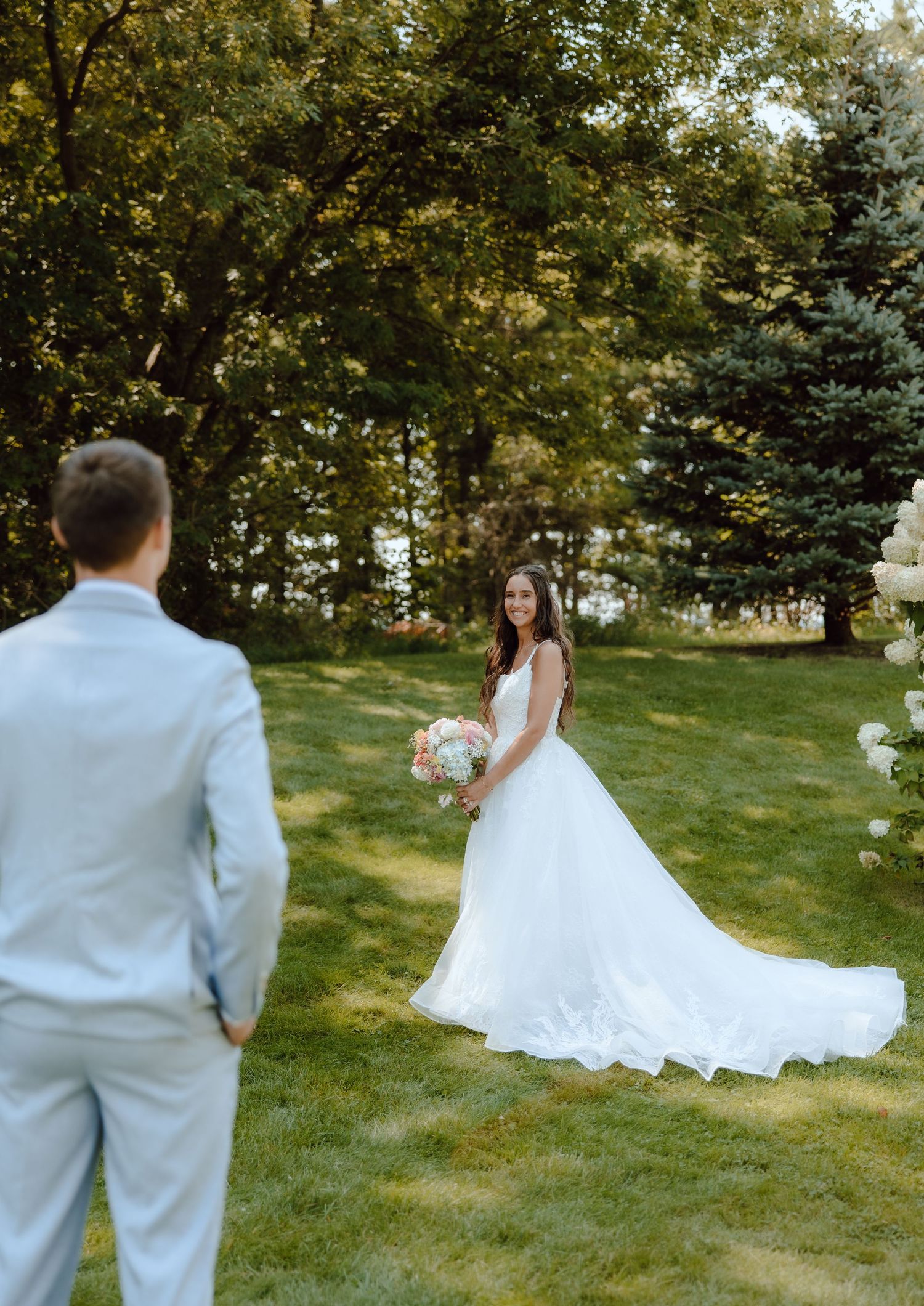 Bride in white gown holds bouquet while groom in light suit approaches during outdoor first look.