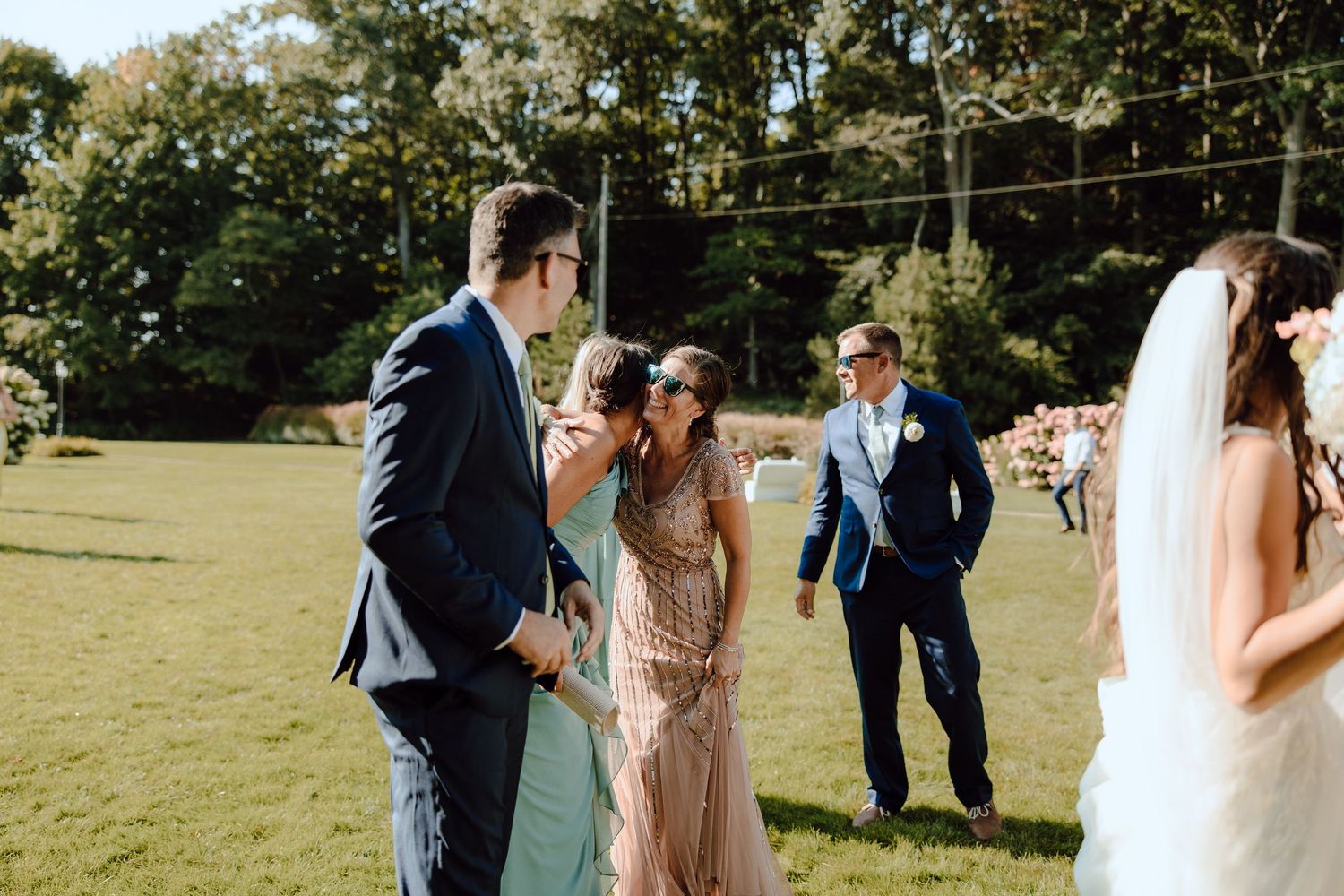 Wedding guests in formal attire greet each other on sunny lawn during outdoor ceremony.