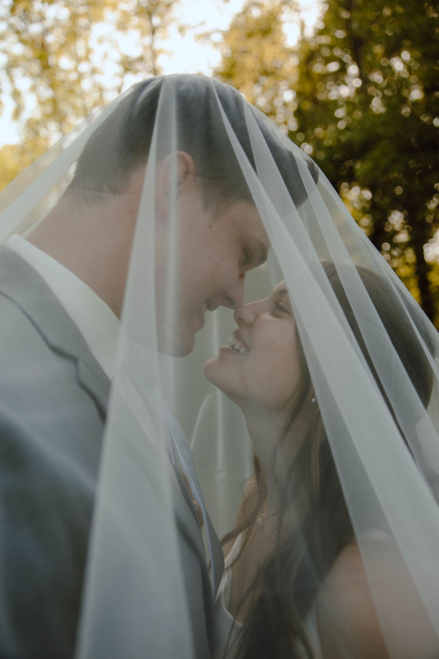 Bride and groom sharing a romantic moment under a flowing white veil with trees in background.