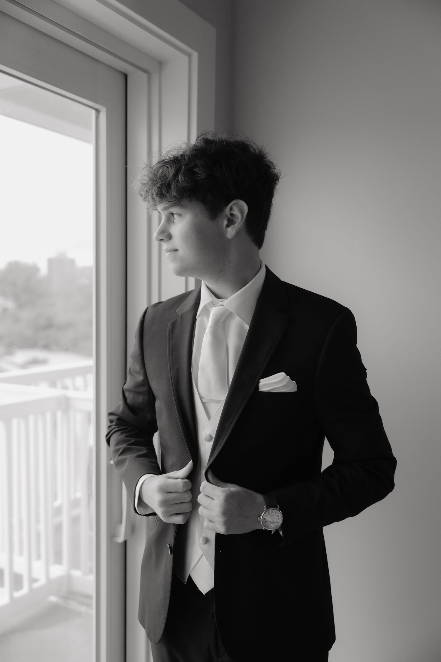 Dapper groom in black tuxedo with white tie gazes out window in black and white portrait.