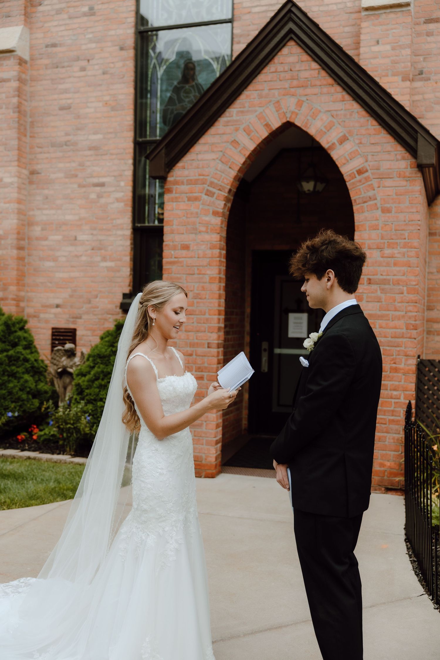 Bride reads vows to groom outside brick church entrance on wedding day.