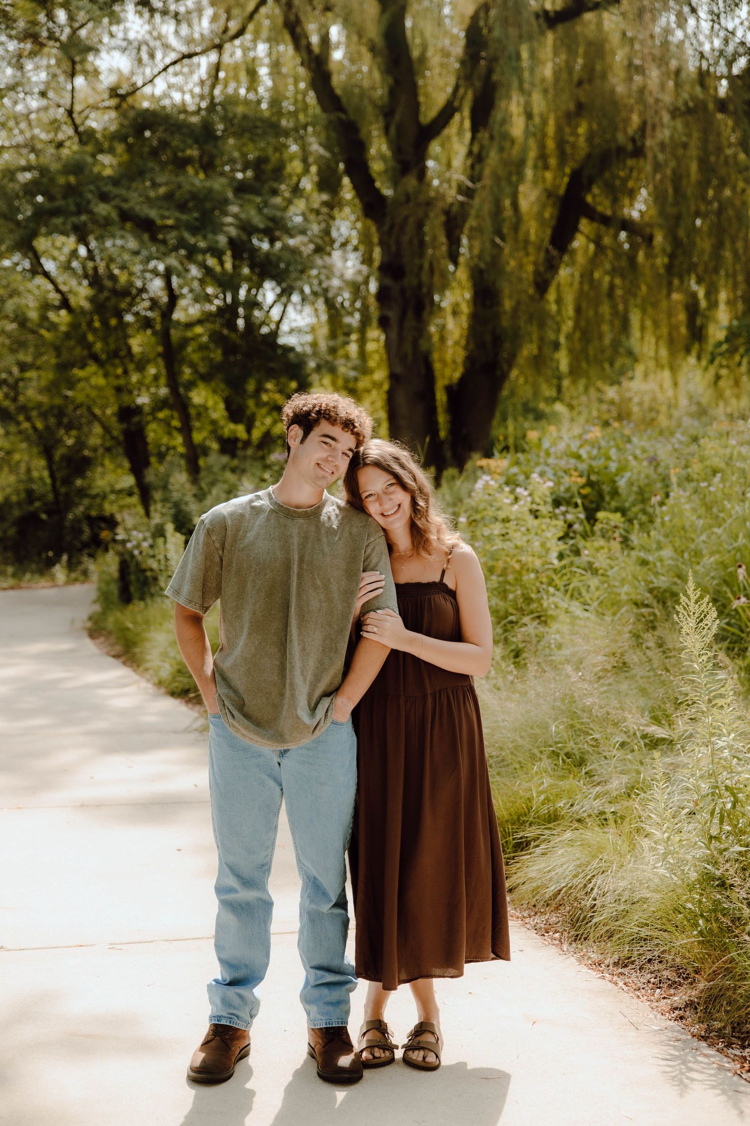 A smiling couple poses on a sunlit garden path surrounded by lush green trees and shrubs.