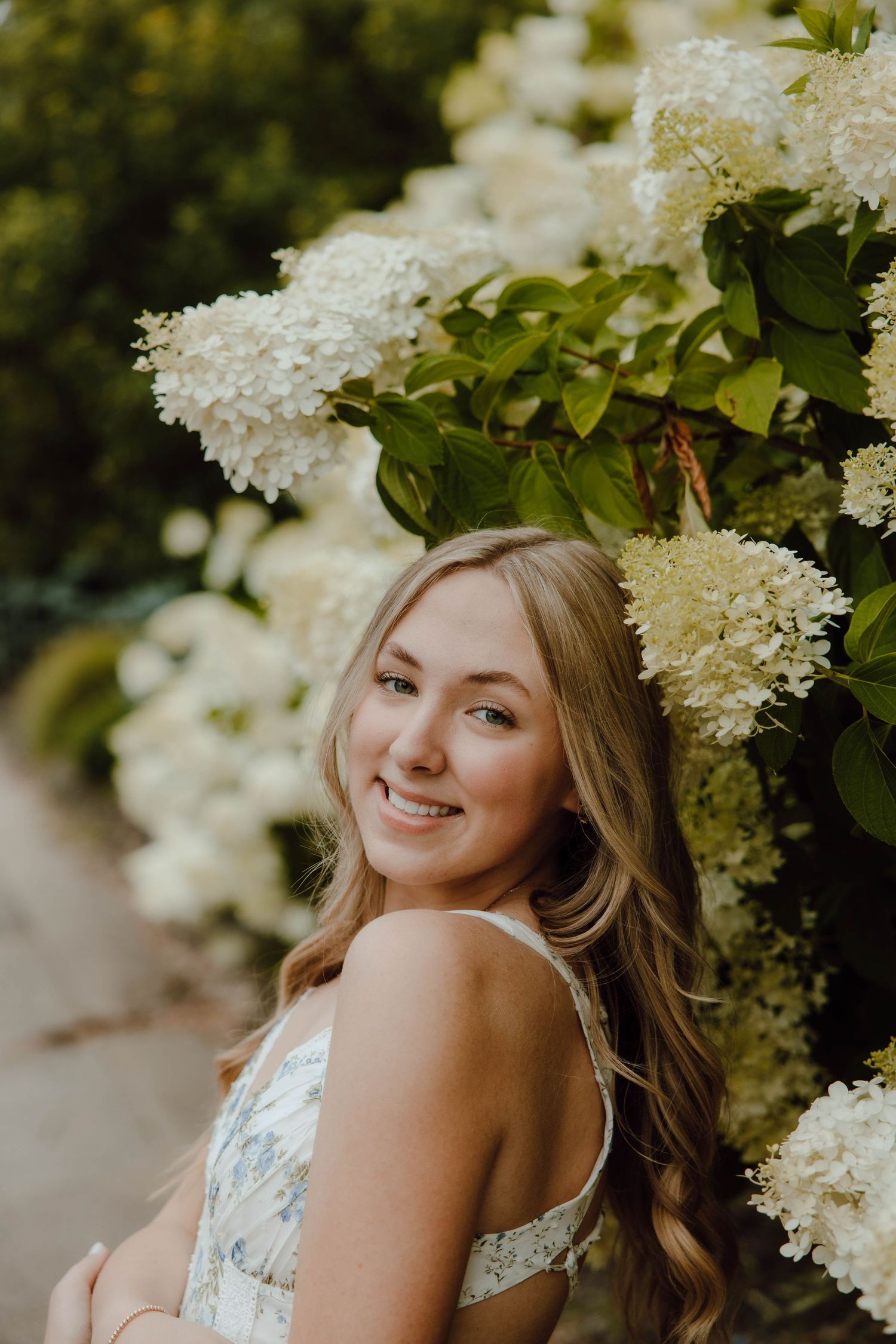 Young woman smiling among white hydrangea blooms in a floral dress, portrait photography outdoors.