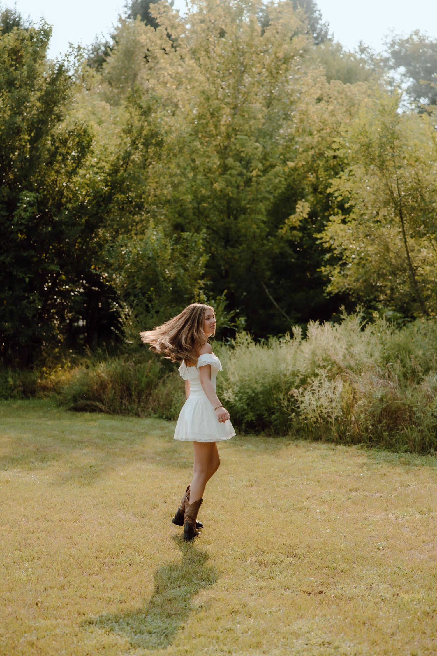 Young woman in white mini dress and cowboy boots twirling in a green meadow surrounded by lush trees.