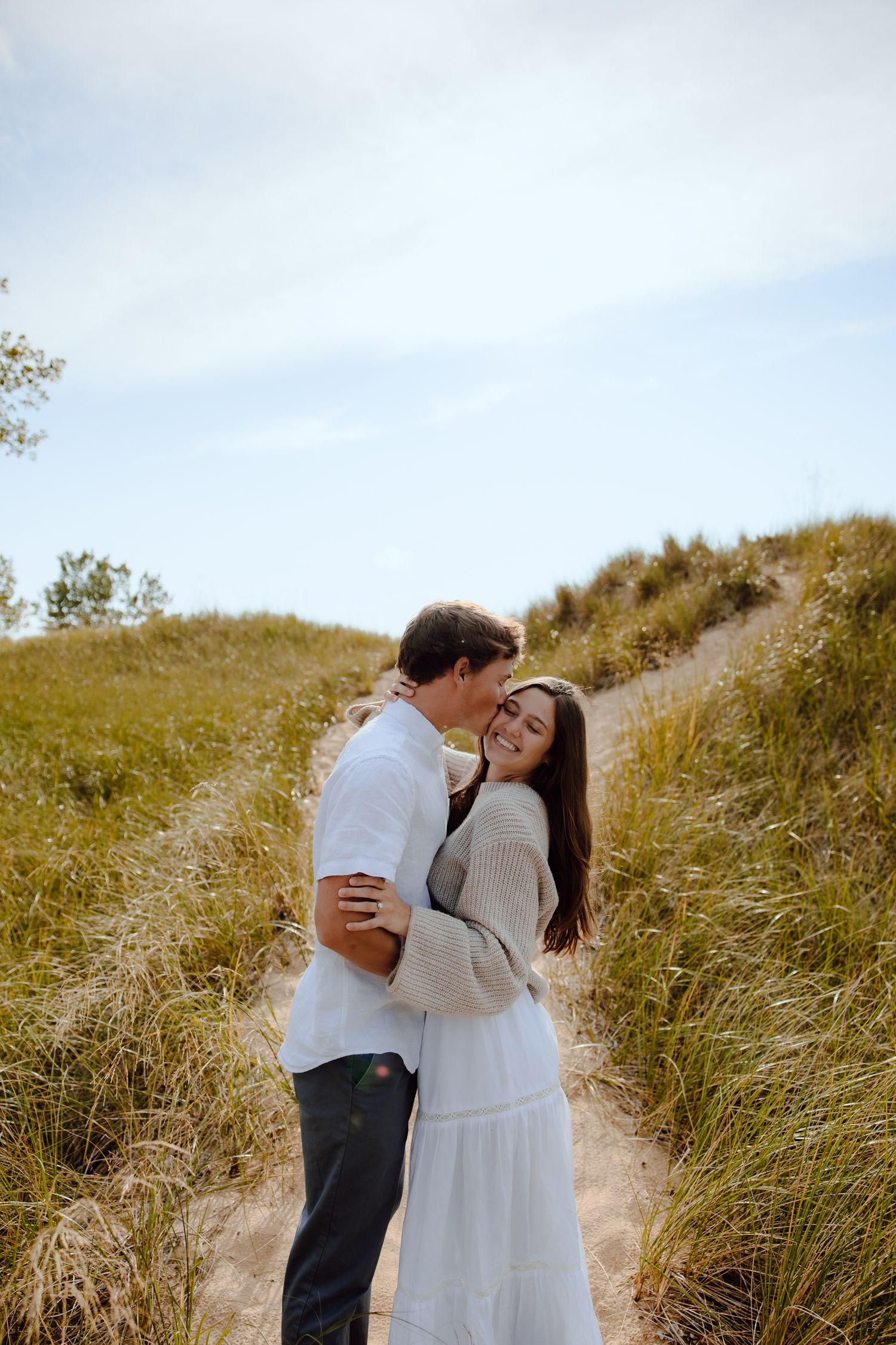 Couple in white and cream outfits embracing on sandy dune path.