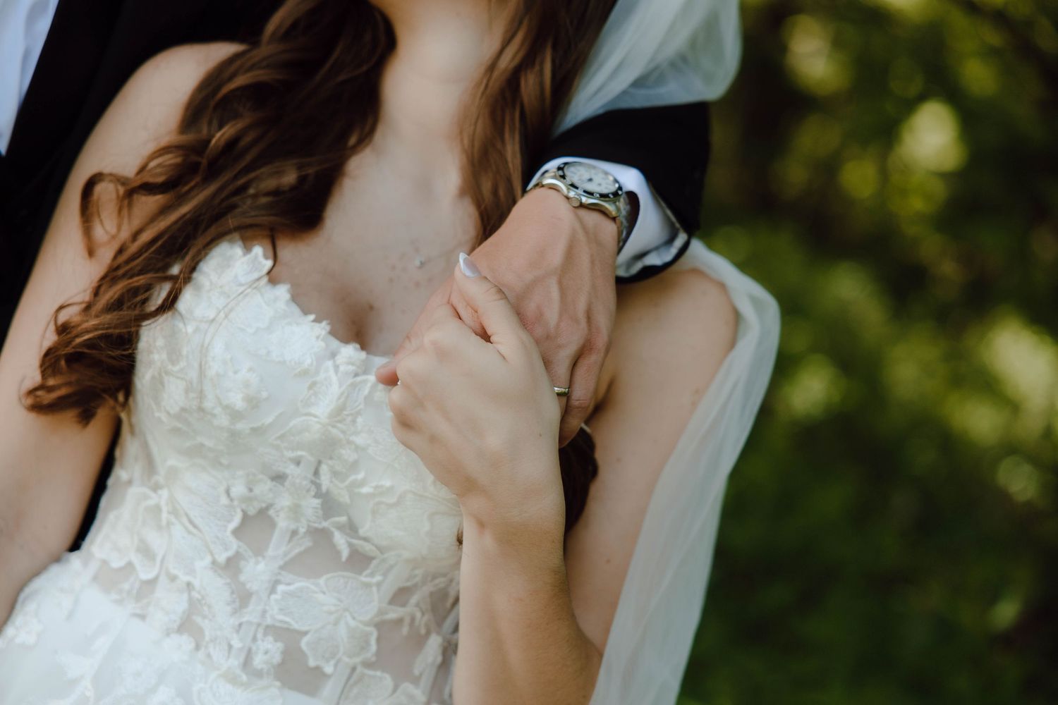Bride and groom holding hands closeup, bride in white lace wedding dress with groom in black suit outdoors.