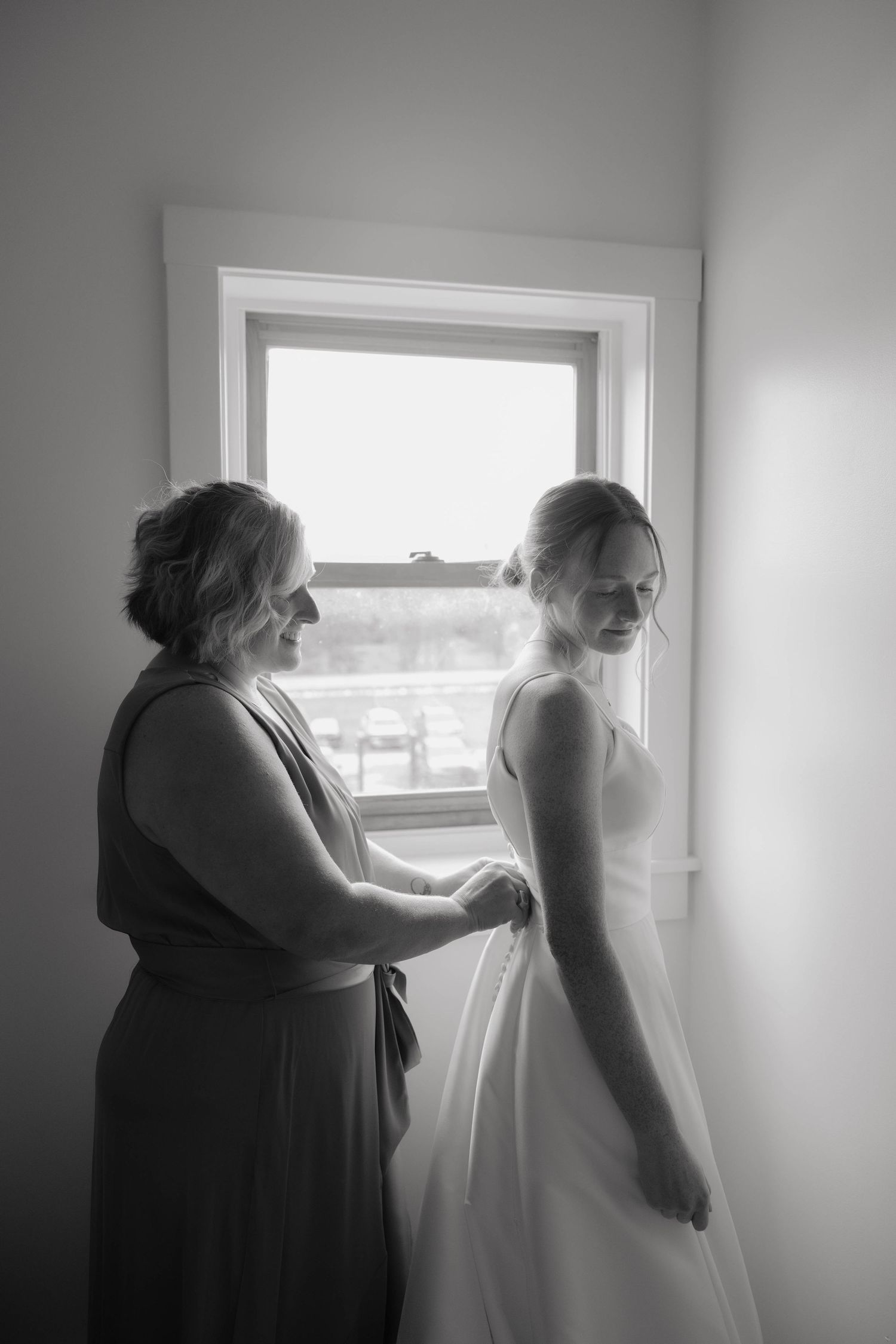 Black and white photo of a bridesmaid helping a bride with her wedding dress near a bright window.