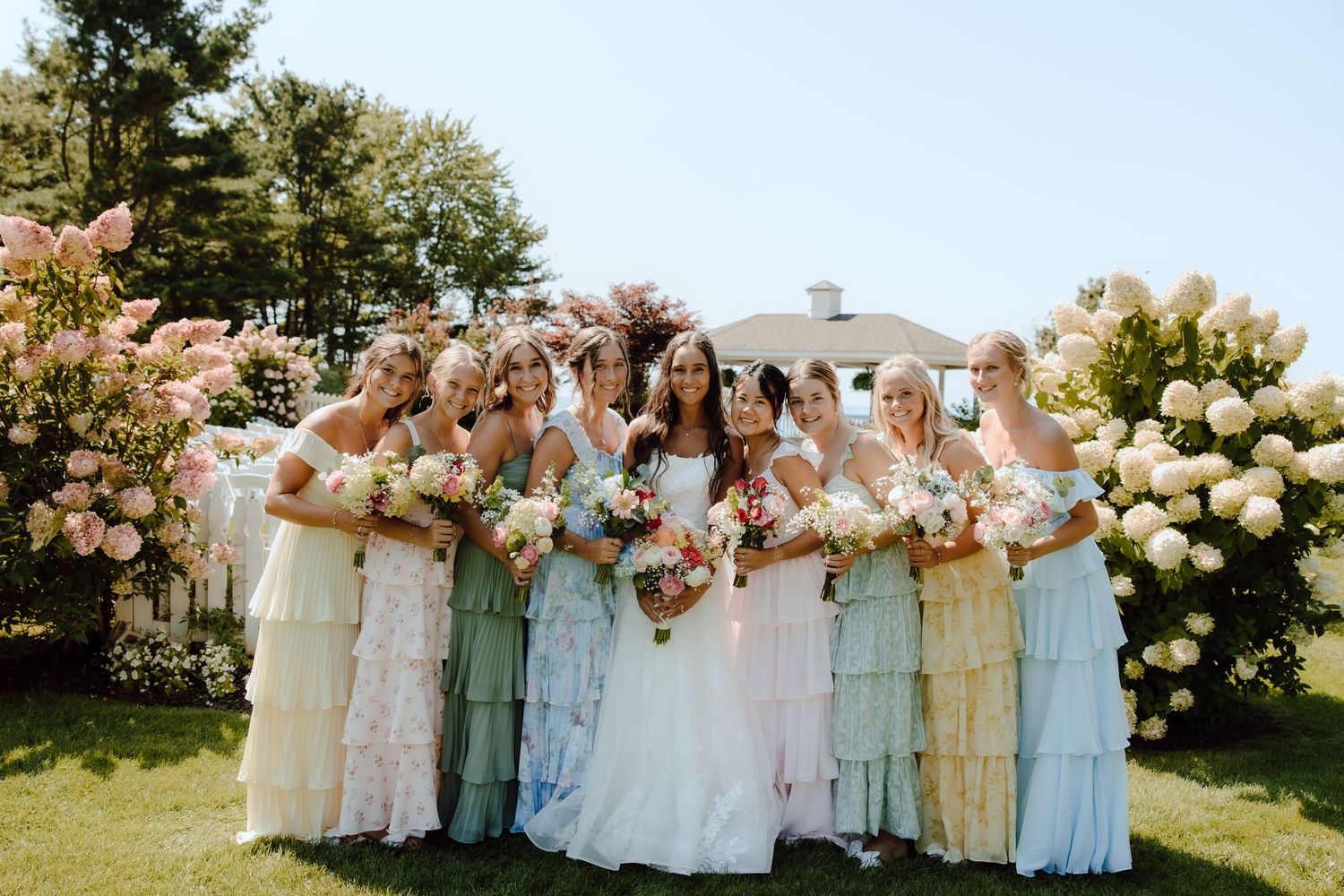Bride and bridesmaids in pastel mismatched dresses holding bouquets outdoors near blooming hydrangeas on a sunny day.