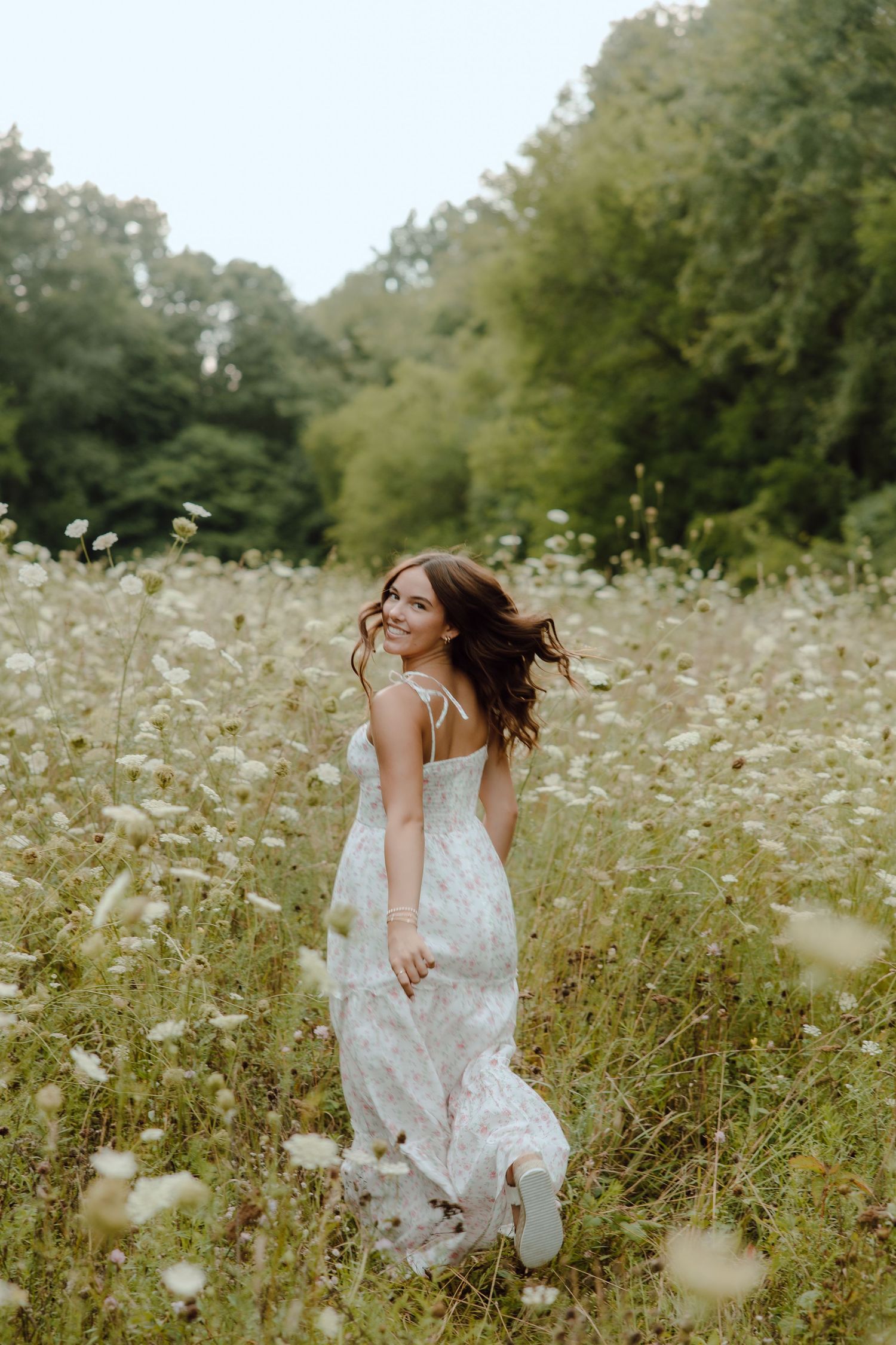Young woman in floral maxi dress smiling over shoulder in wildflower meadow with lush green trees background.