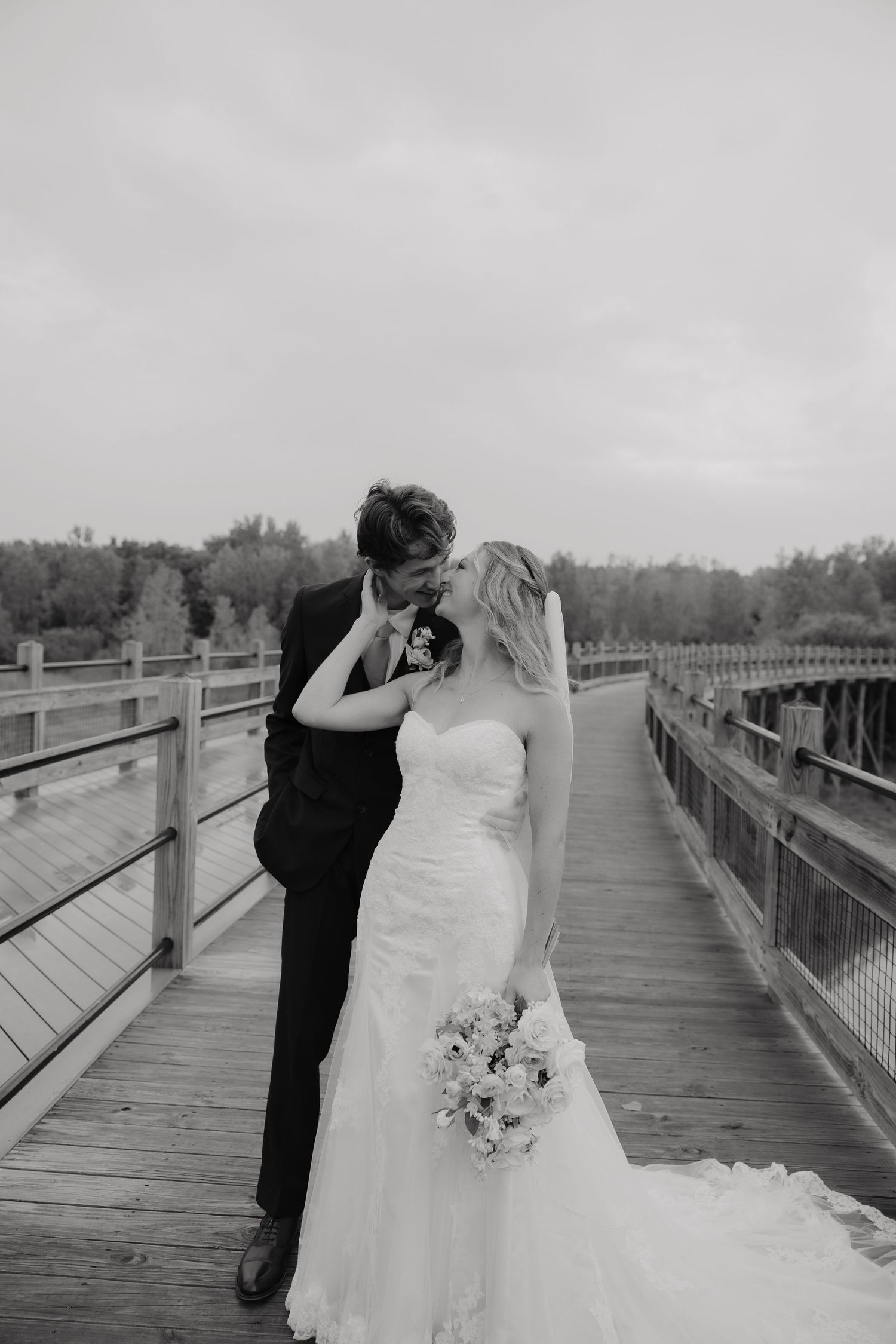 Black and white wedding photo of a bride and groom sharing a kiss on a wooden bridge, bride holding a bouquet.