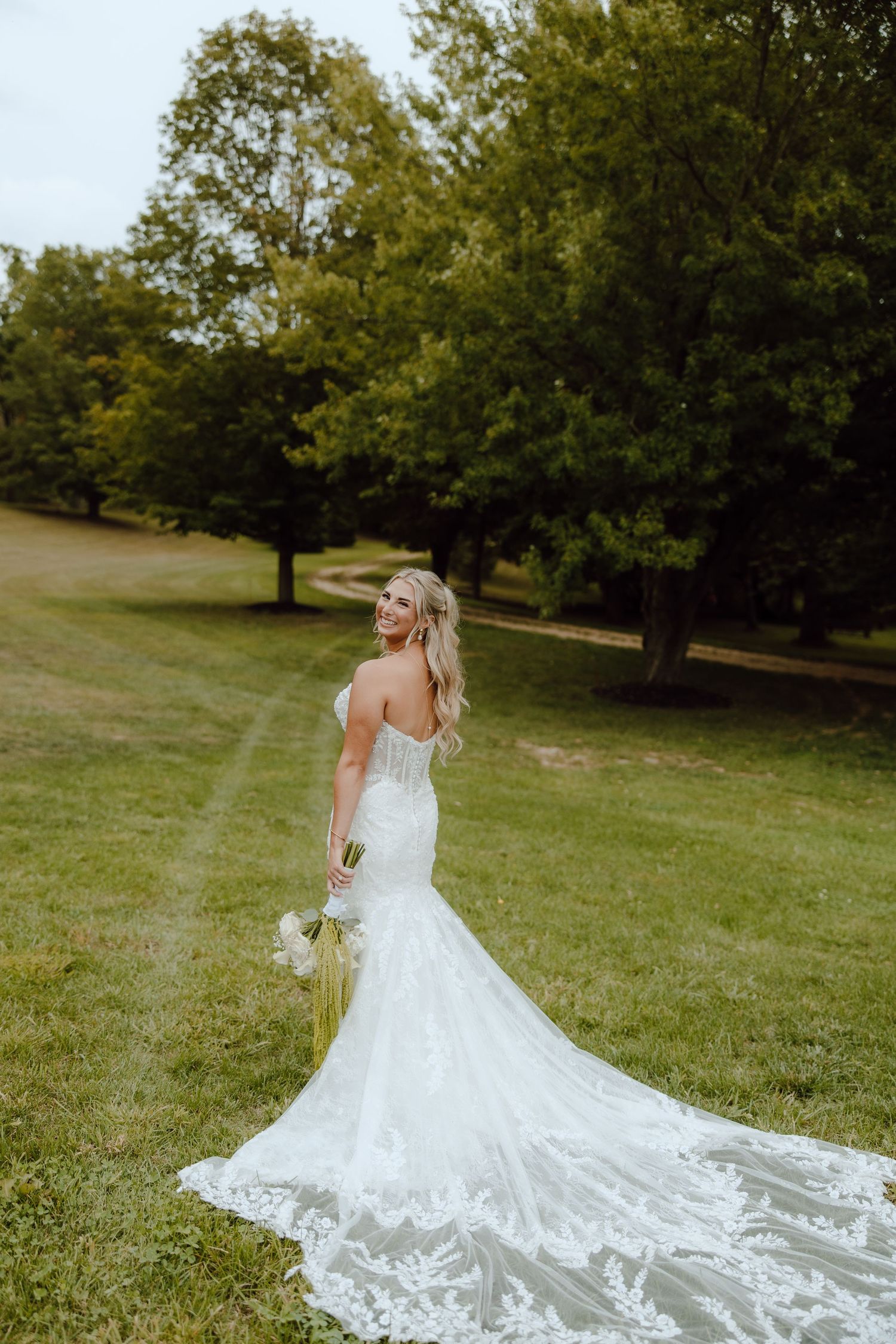 Bride in a strapless lace mermaid wedding gown with a long train holds a bouquet on a green lawn.