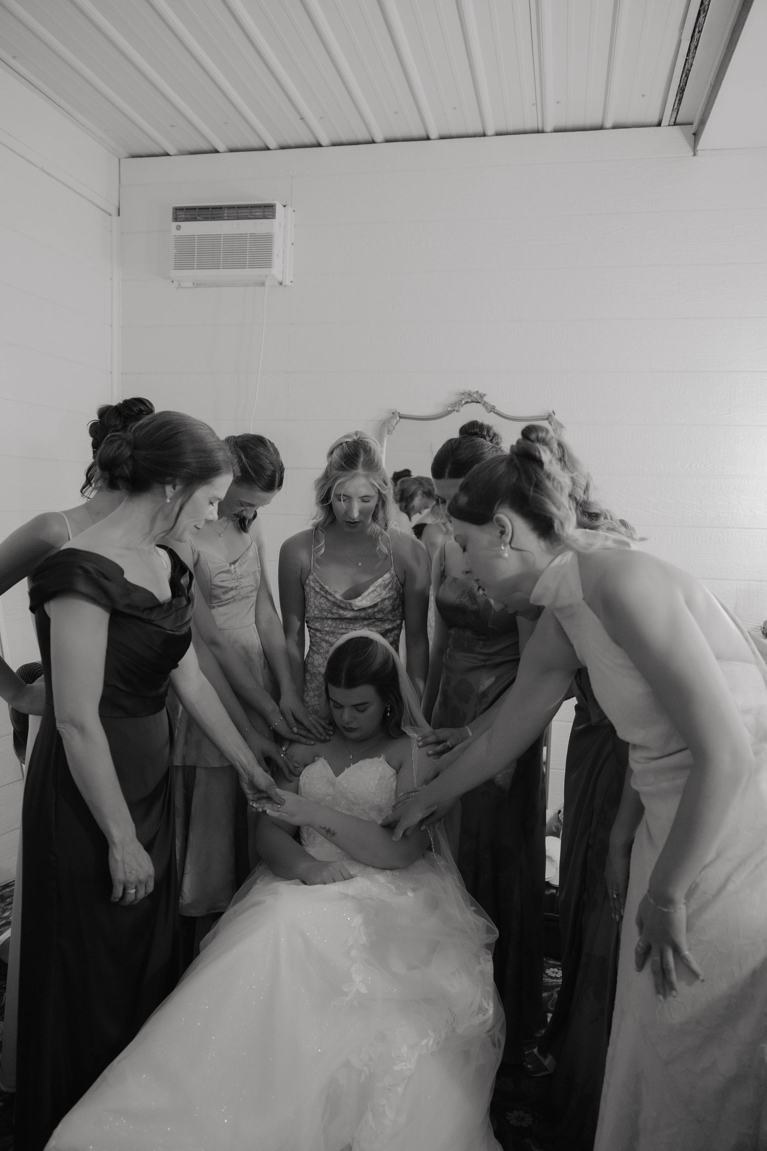 Bridesmaids gather around bride in circle prayer before wedding ceremony in black and white photo.