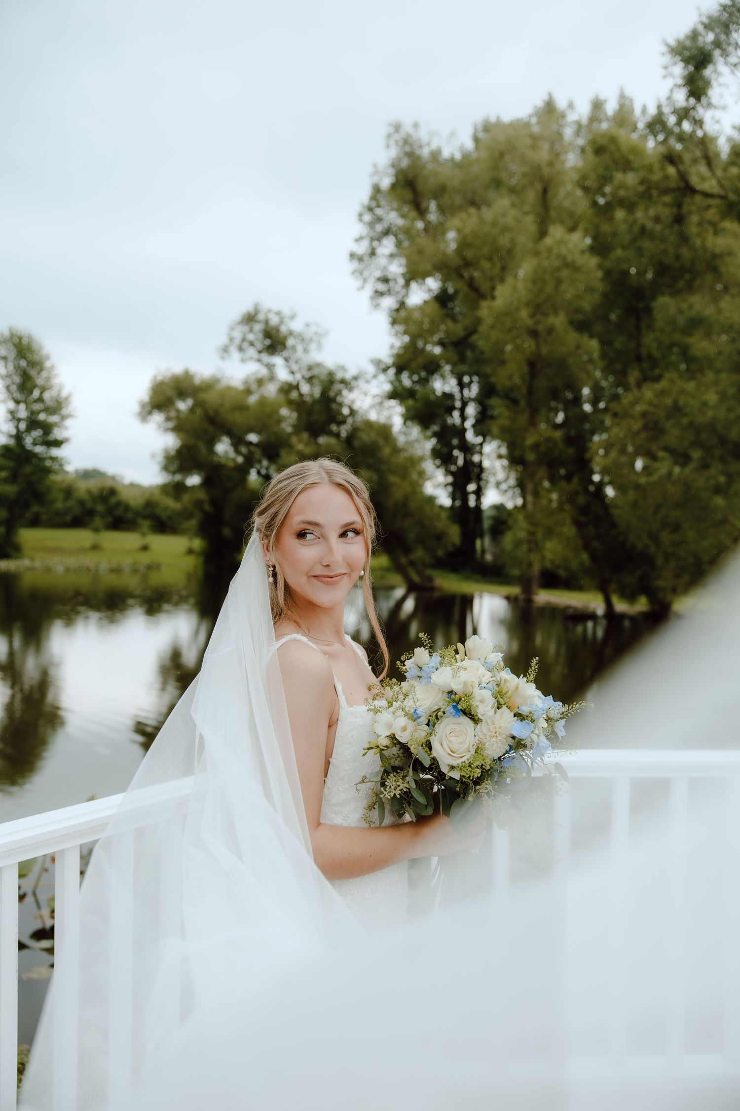 Smiling bride in white wedding dress and veil holding a blue and white bouquet on a bridge overlooking a peaceful pond.