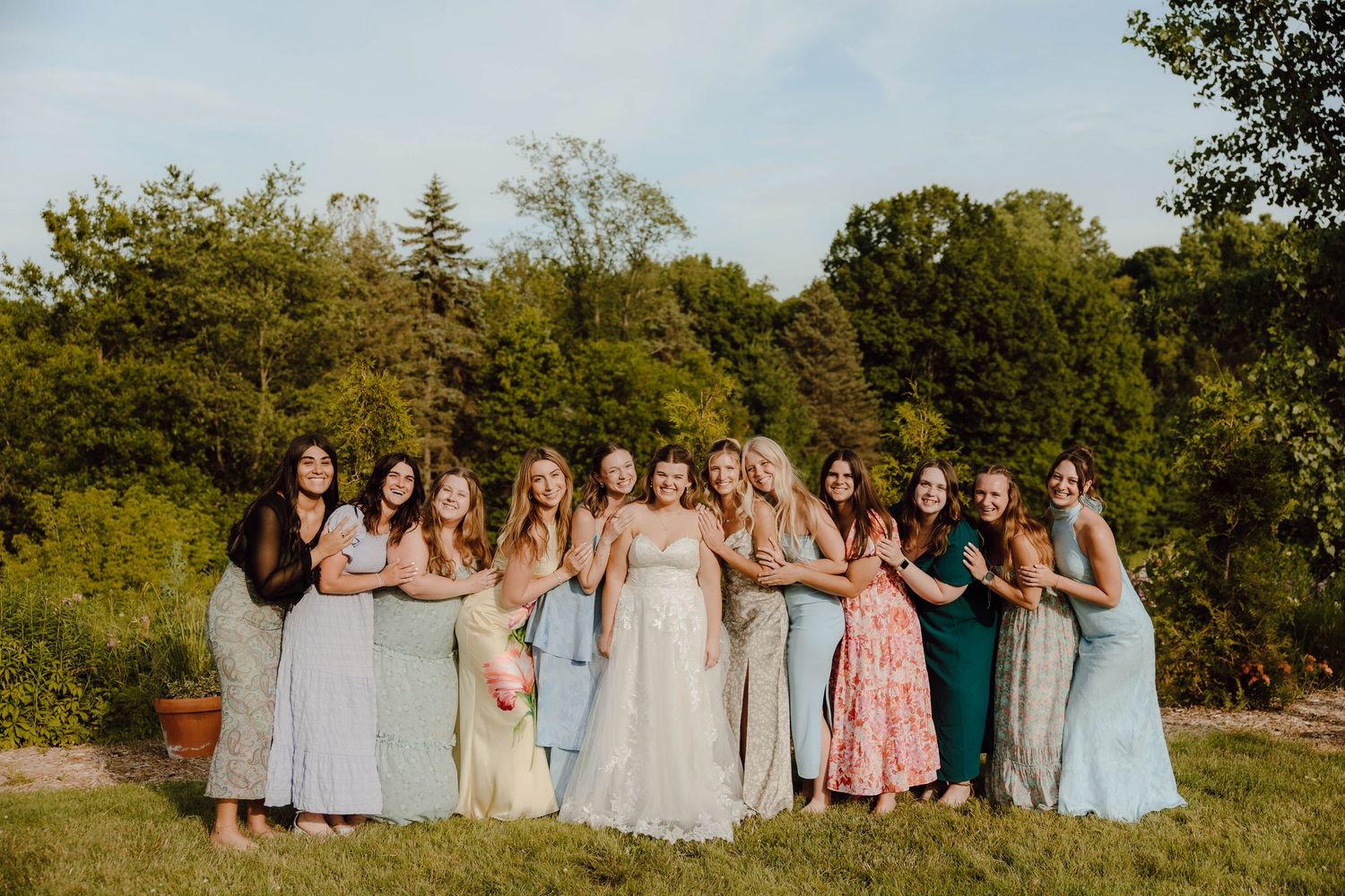 Bride poses with bridesmaids in mismatched colorful gowns on a sunny green lawn.