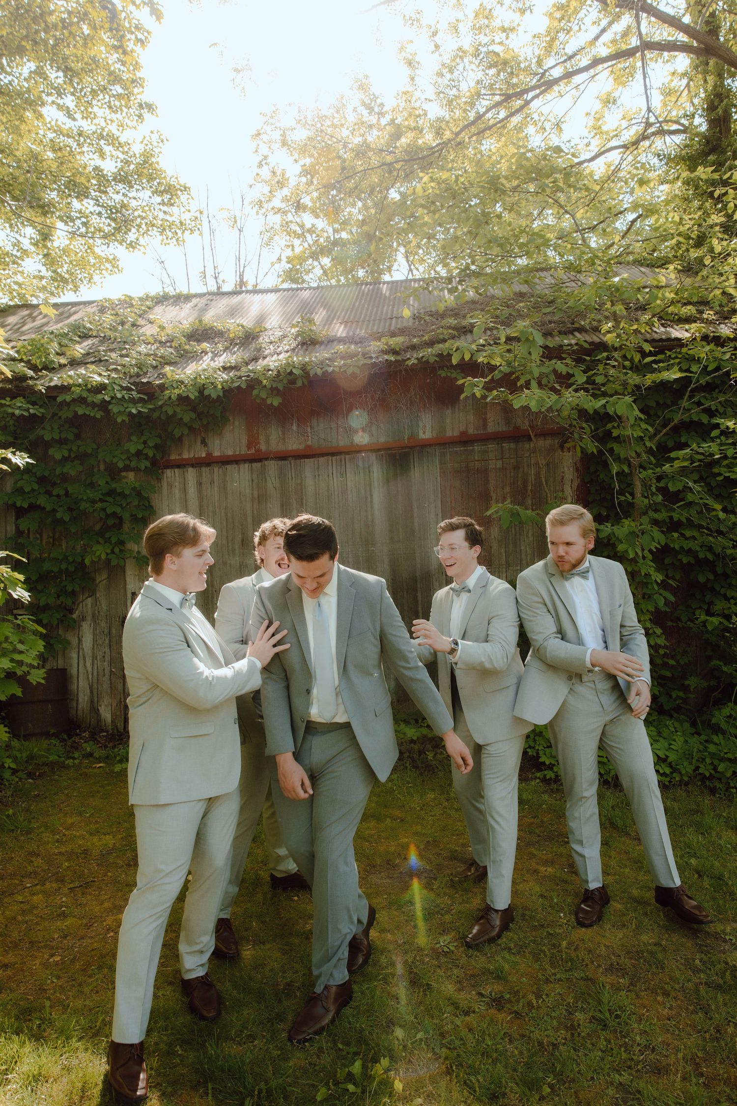 Groom and groomsmen in matching light gray suits laugh together in front of a rustic wooden barn.