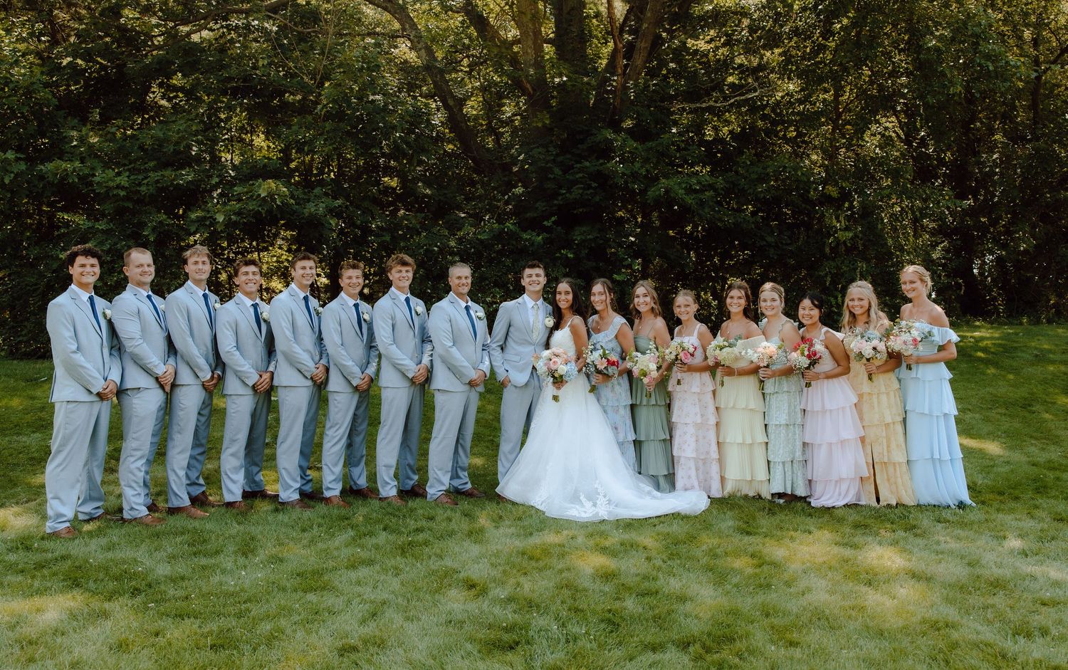 Wedding party posing on grass with groomsmen in gray suits and bridesmaids in pastel dresses.