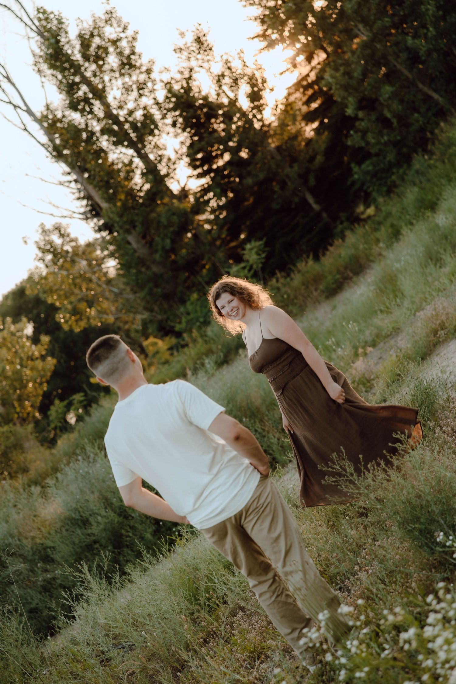 Couple walking in a sunlit meadow, woman in brown dress smiling, man in white tee, surrounded by lush greenery.