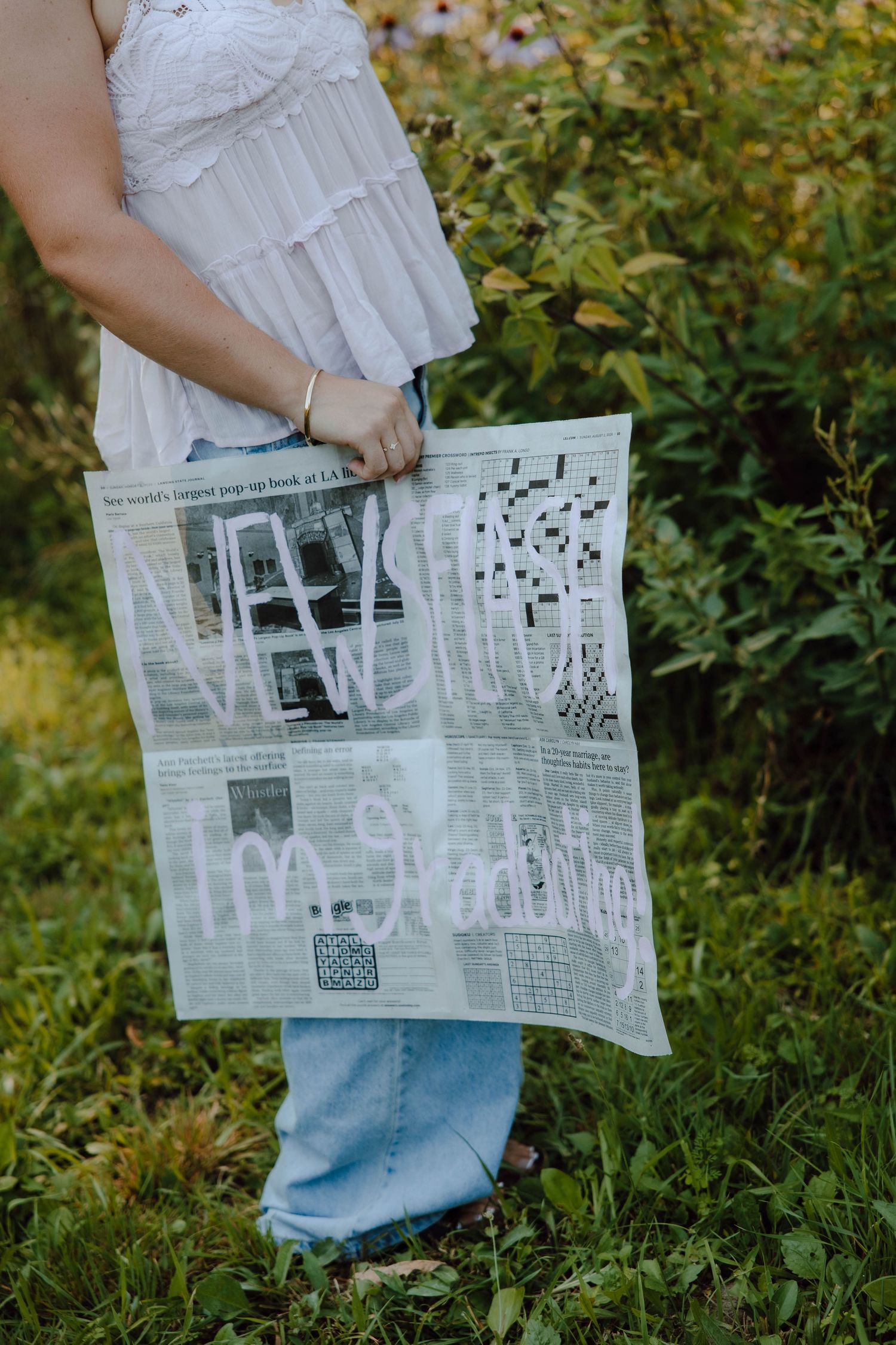 Person holding a newspaper with handwritten text saying I'm graduating outdoors near green bushes.