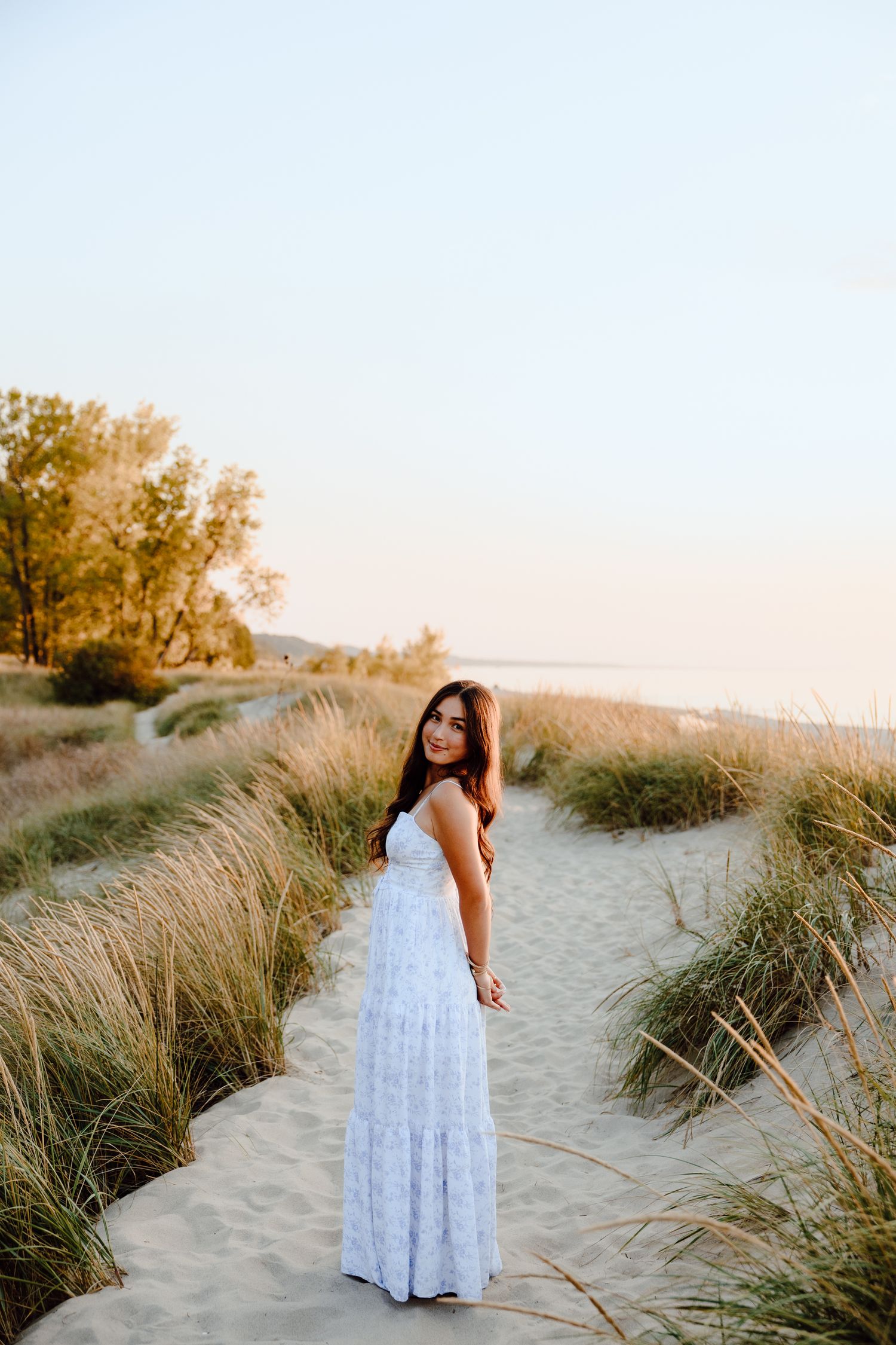 Person in white maxi dress poses among beach grass during sunset.