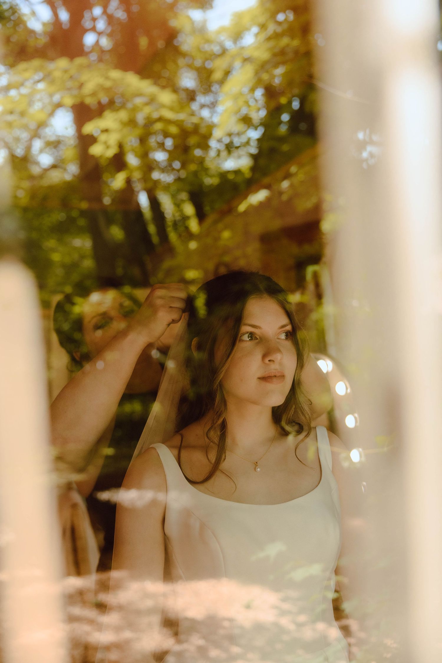Bride having her hair styled outdoors, captured through a sunlit window surrounded by lush green trees.
