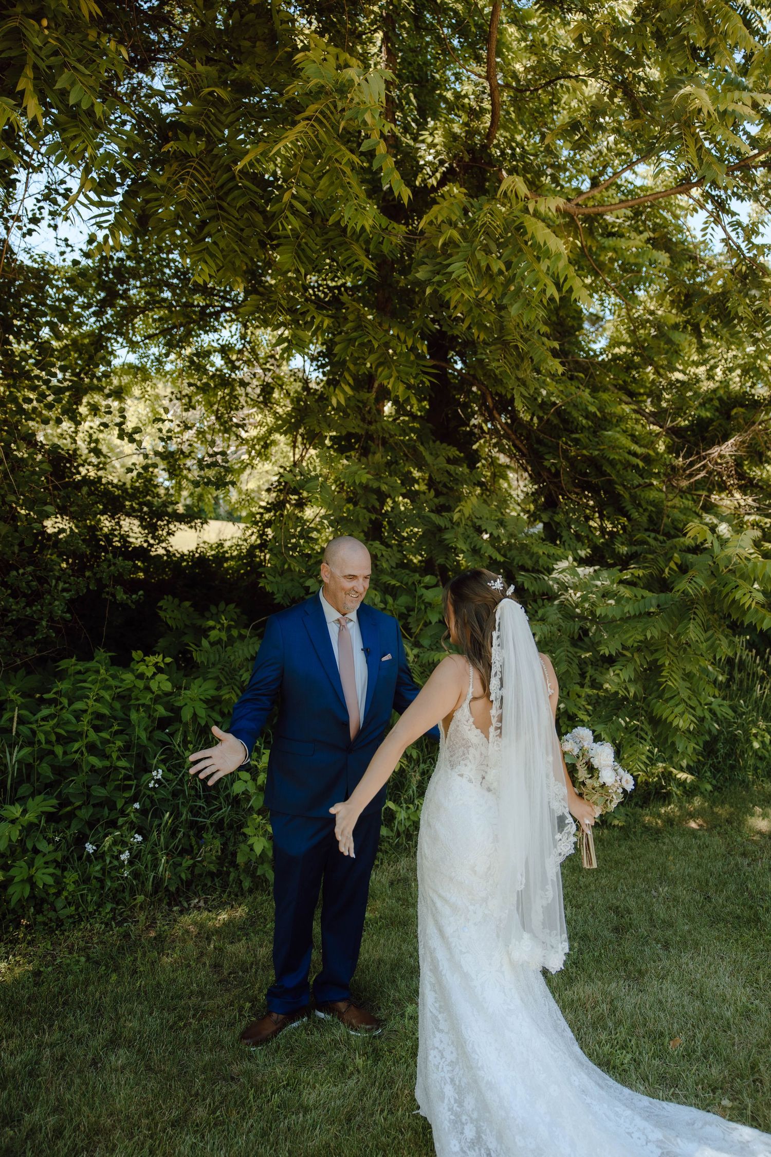 Bride in lace gown with veil holds groom's hand during outdoor first look moment in lush green garden.
