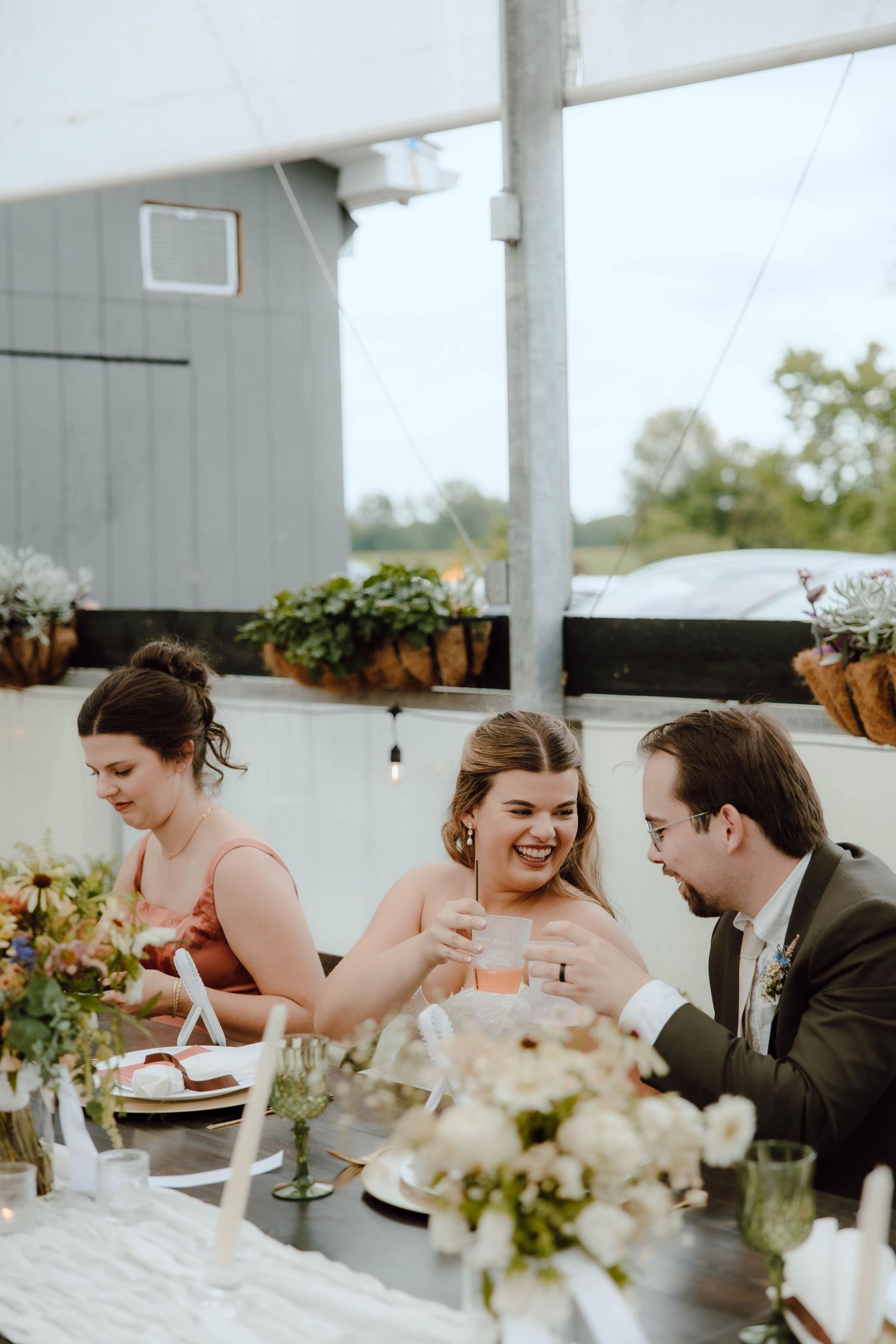 Bride and groom clink glasses with bridesmaid at elegant outdoor wedding reception under white tent.