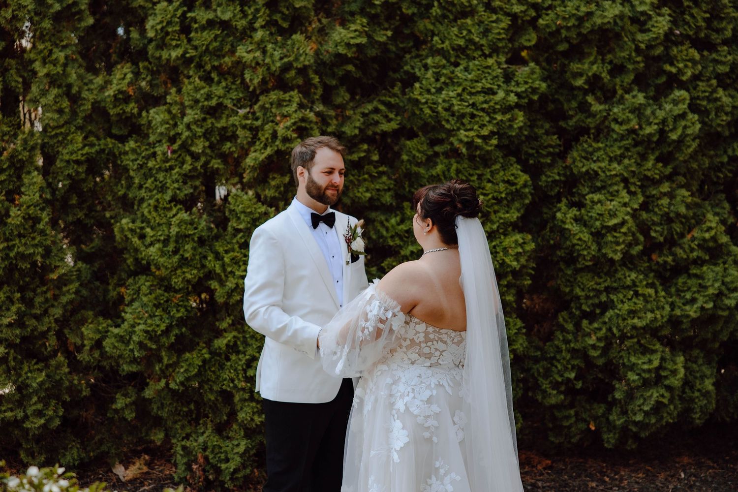 A romantic wedding moment captured against lush green hedges with the couple in elegant white attire.