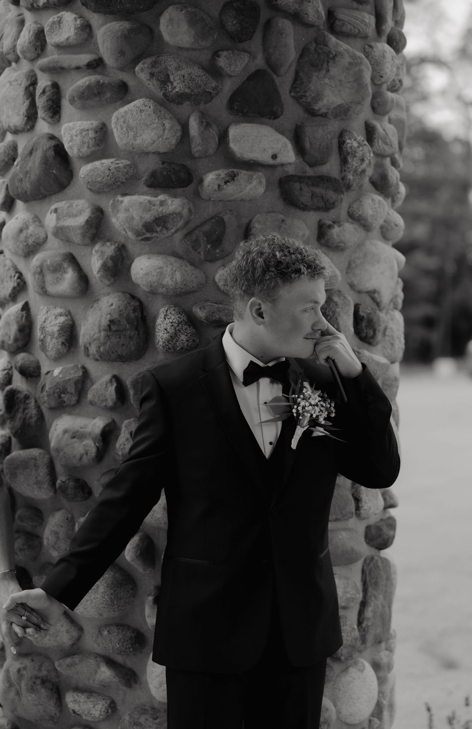 Groom in black tuxedo with boutonniere stands by stone pillar, emotional on wedding day.