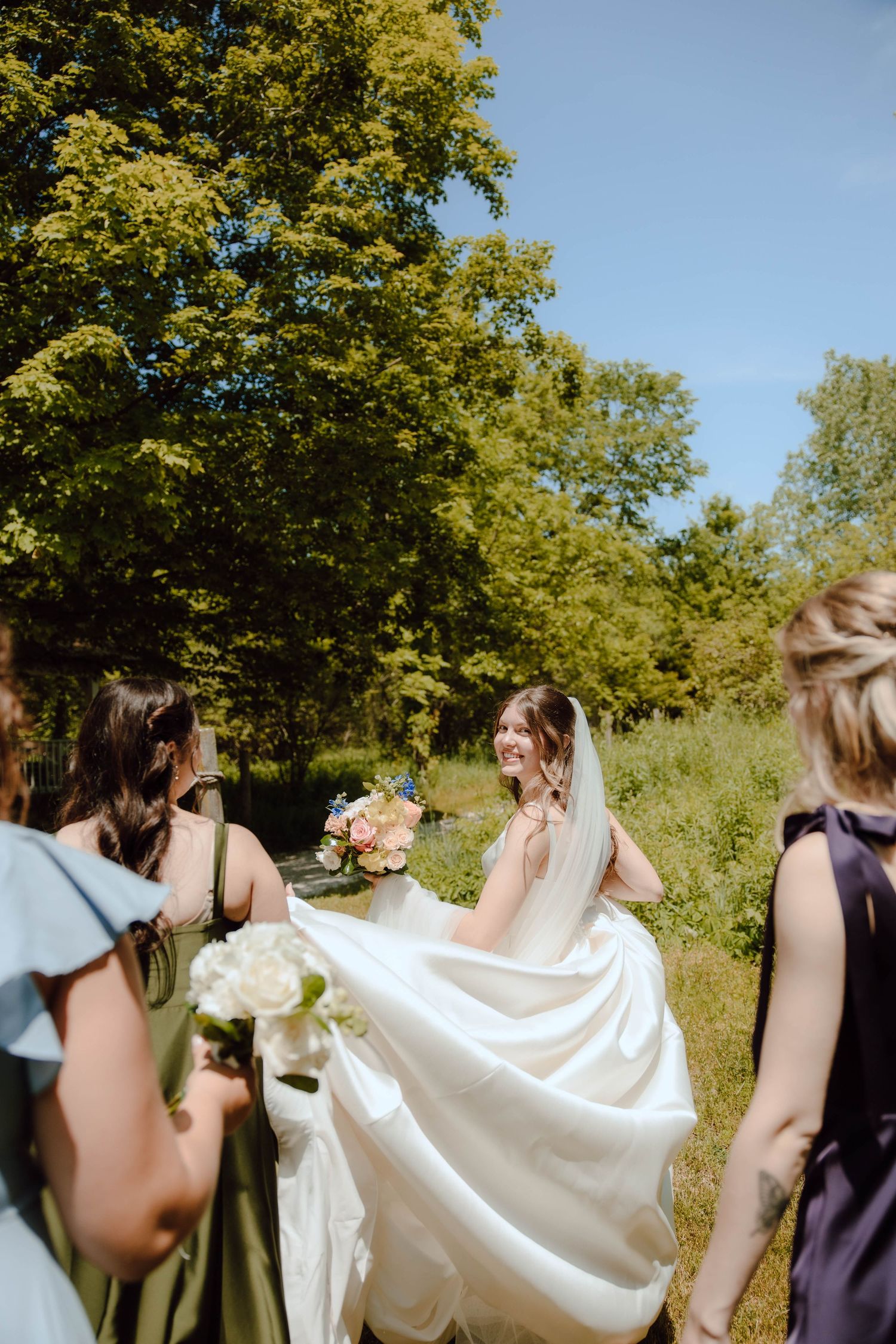 Bride in white gown laughs with bridesmaids in green and purple dresses outdoors on a sunny day.