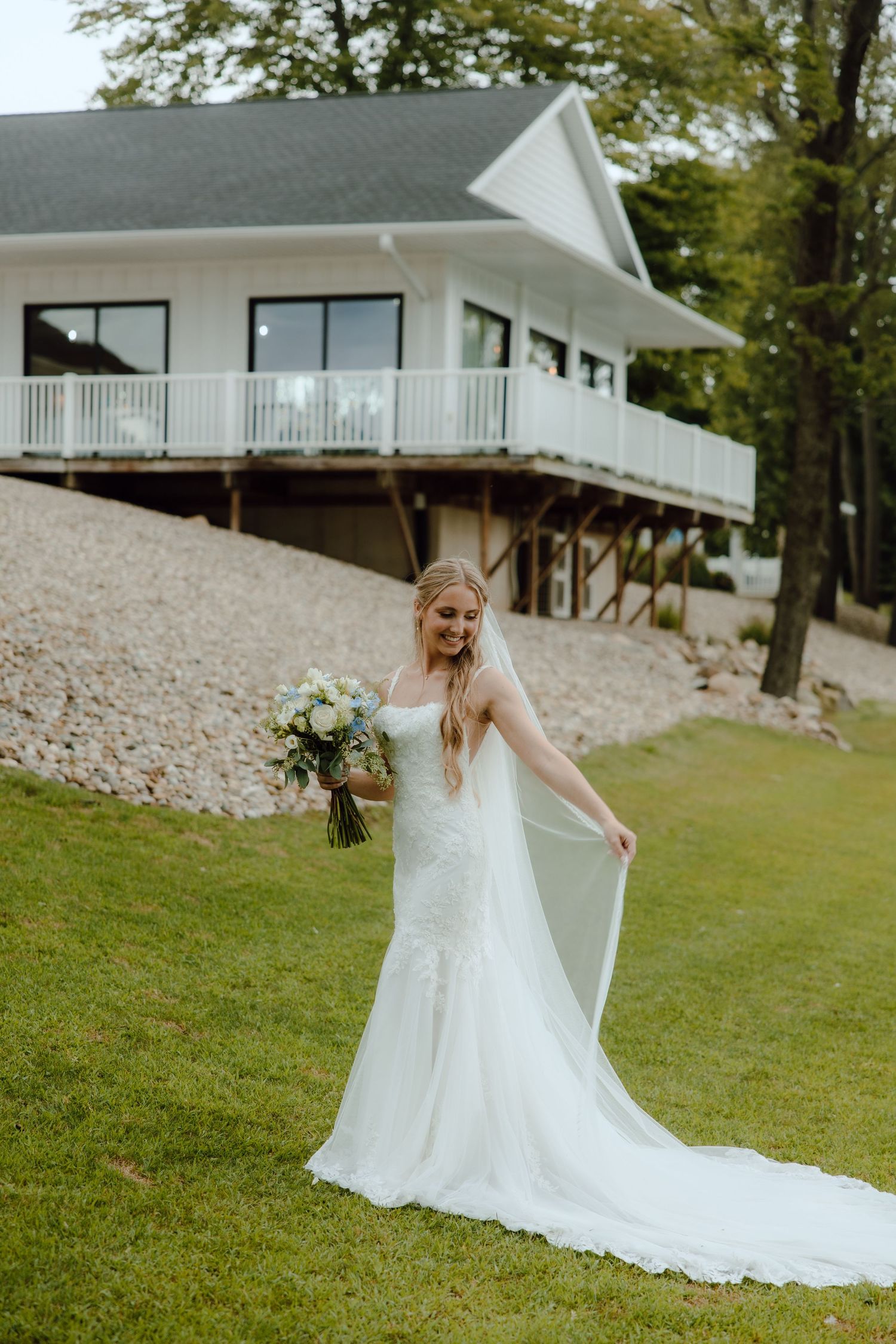Bride in white mermaid gown holding blue and white bouquet standing on green lawn before elegant white venue.