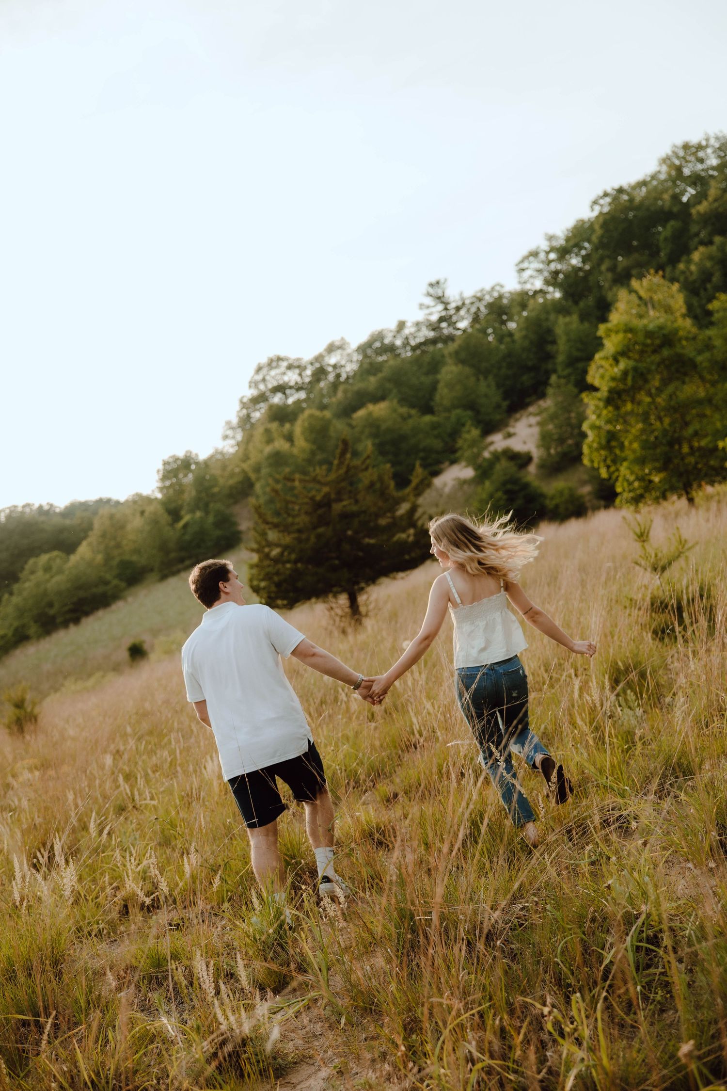 A couple holds hands while walking through a golden meadow with lush green trees in the background.
