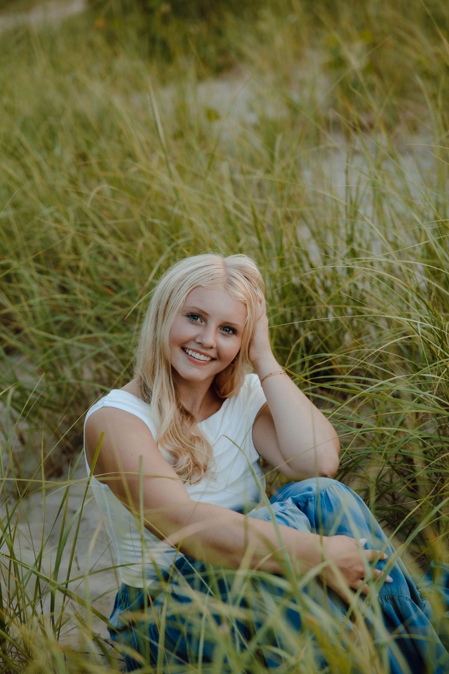 Smiling blonde teen girl sitting in tall beach grass wearing white top and jeans, posing for senior portrait.