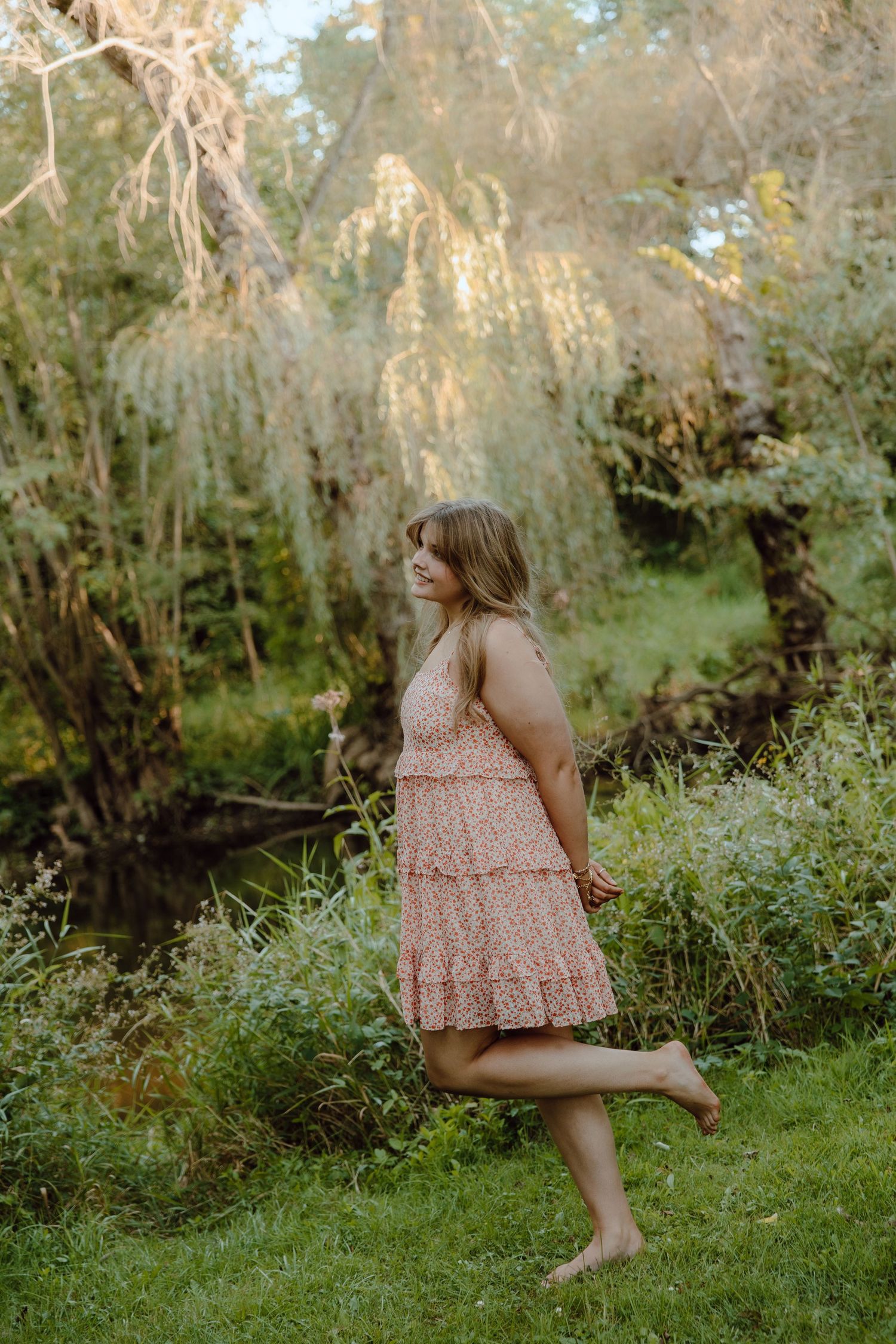 Young woman in a pink floral sundress walking barefoot on green grass near a weeping willow tree.