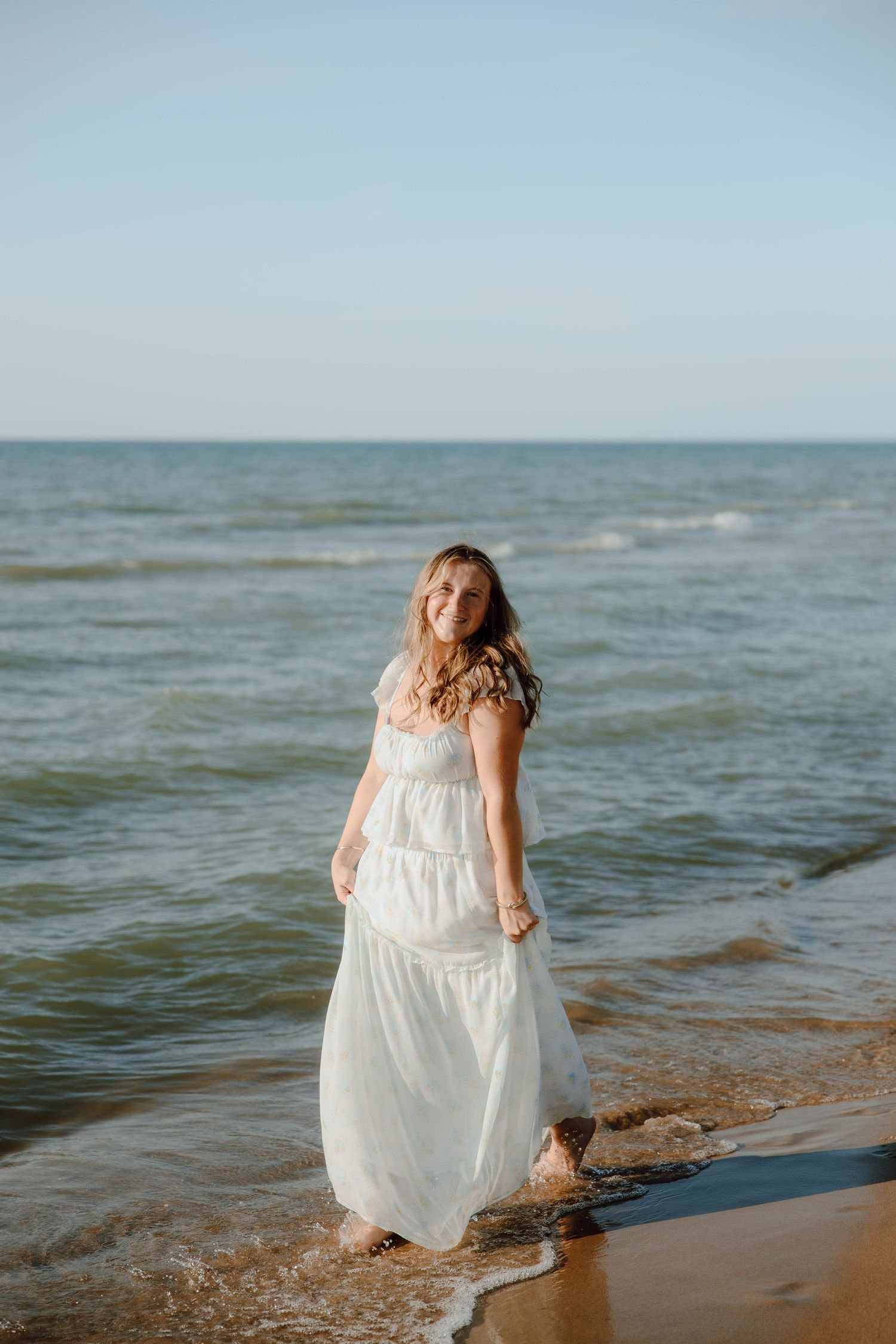 Smiling woman in white maxi dress standing barefoot in ocean waves on sandy beach.