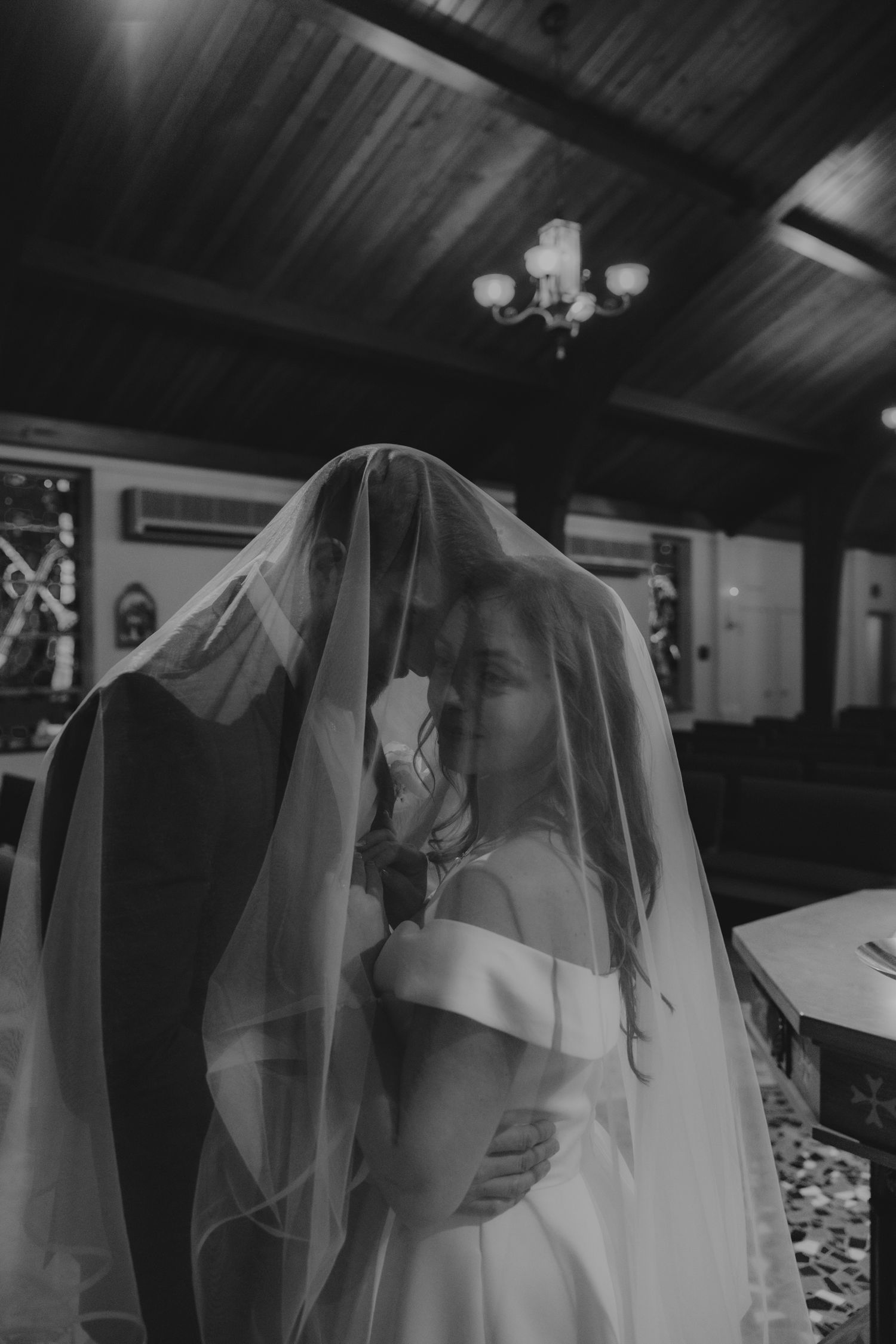 A dramatic black and white photograph of a bride wearing a flowing veil in a dimly lit indoor setting.
