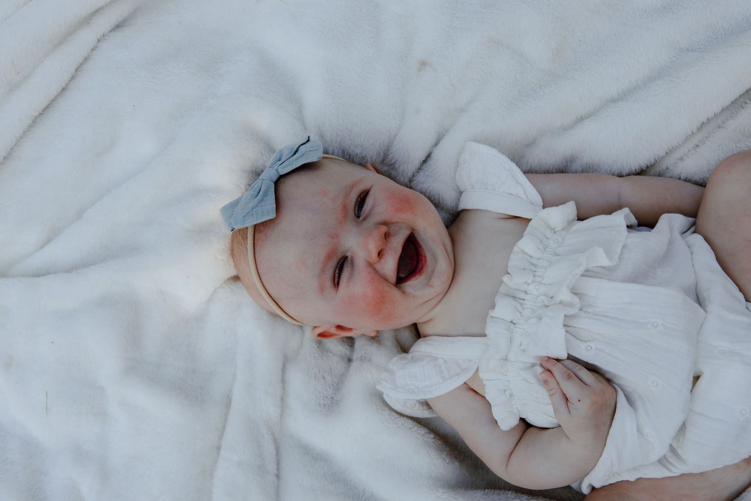 Baby wearing white outfit and blue bow laughing on white blanket.