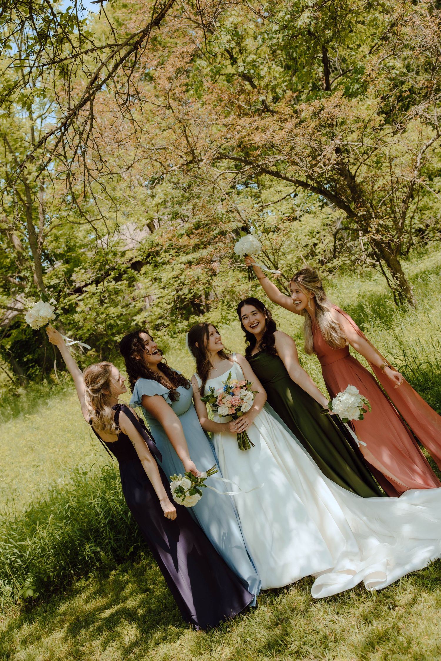 Bride and four bridesmaids in colorful mismatched gowns celebrate joyfully under trees raising bouquets.