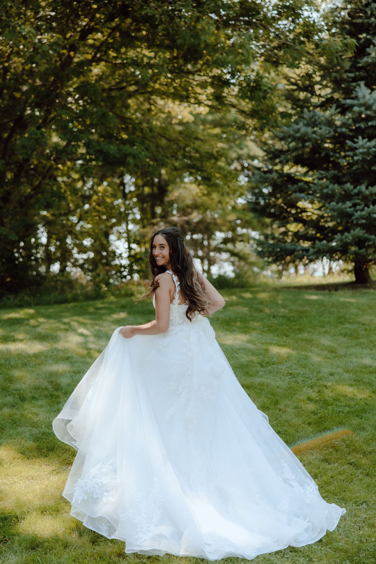 Bride in a flowing white ball gown twirling outdoors on green grass with lush trees in the background.
