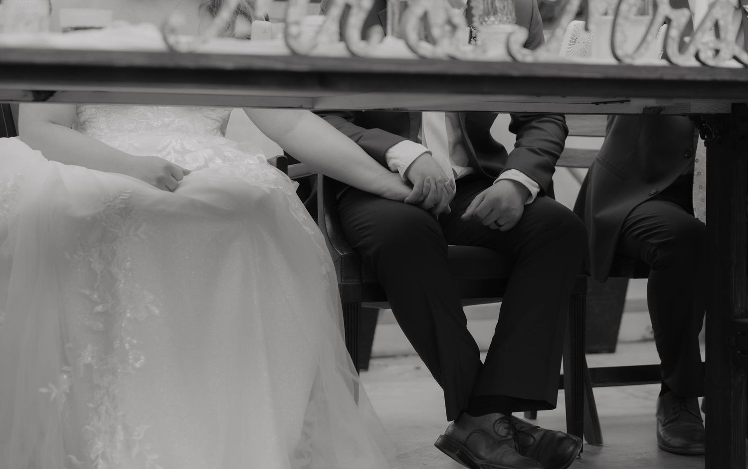 Black and white wedding photo of bride and groom holding hands while seated at a table beneath a Mr. and Mrs. sign.