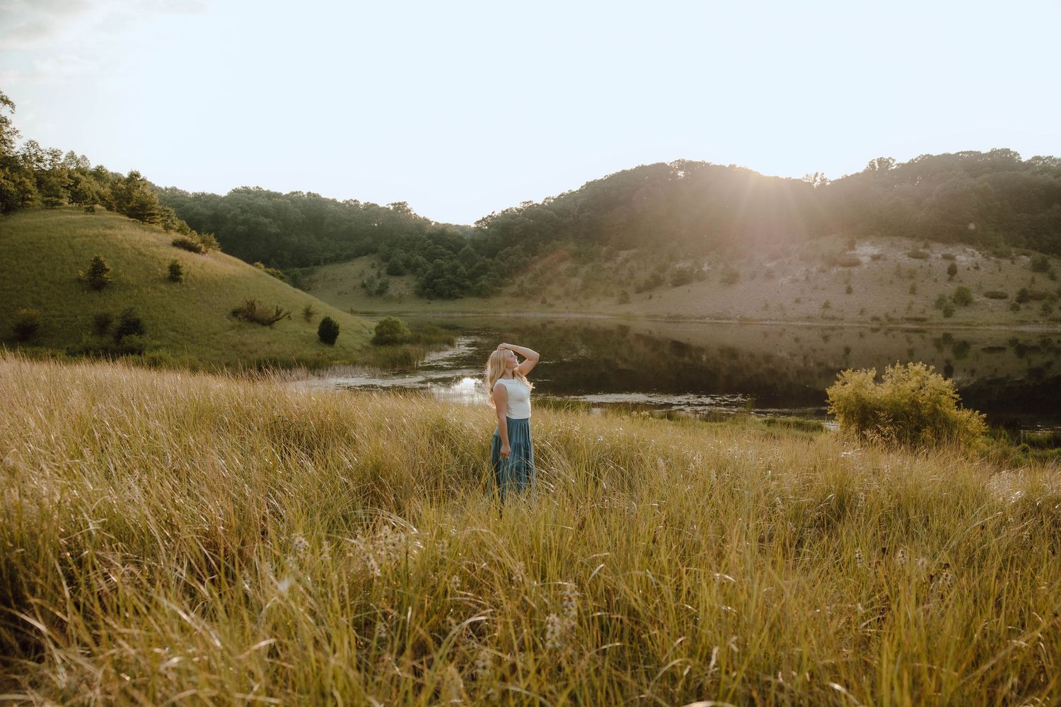 Person standing in golden meadow with rolling green hills and serene pond at sunset.