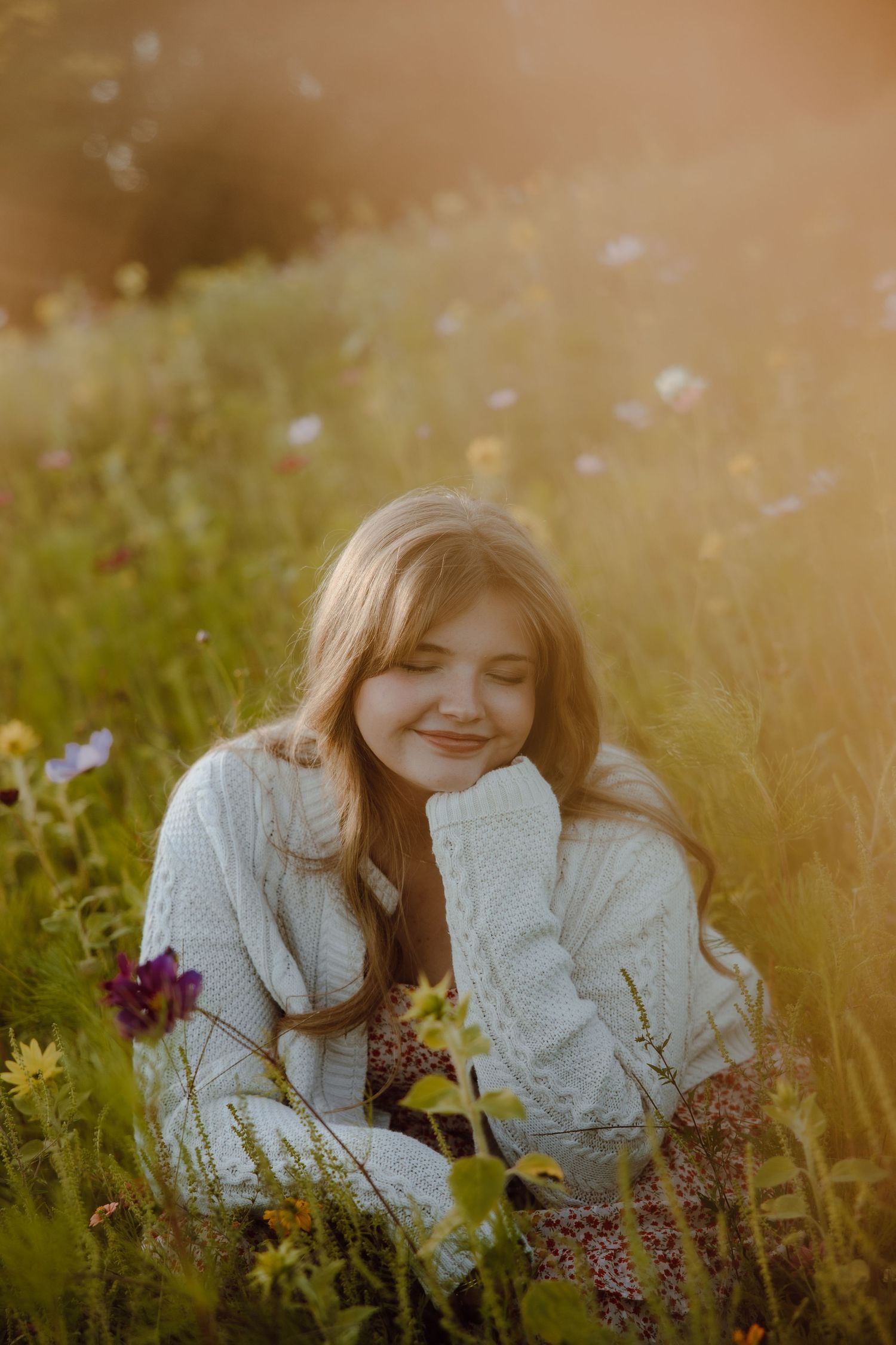 Young girl with long blonde hair sits smiling in a sunlit wildflower meadow wearing a white knit sweater.