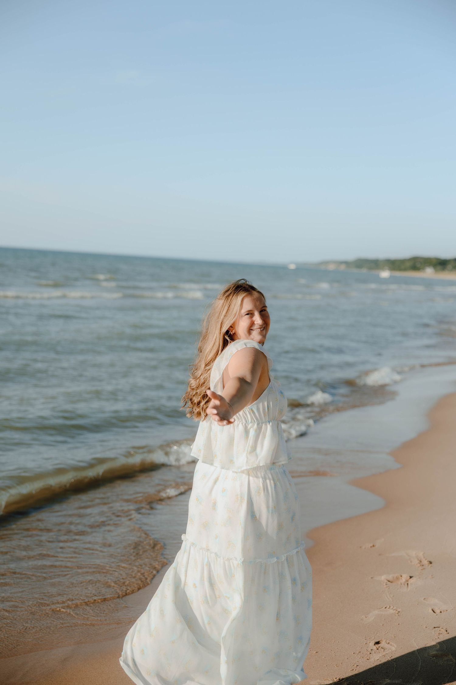 A smiling woman in a white maxi dress walks along a sandy beach shoreline with ocean waves in the background.