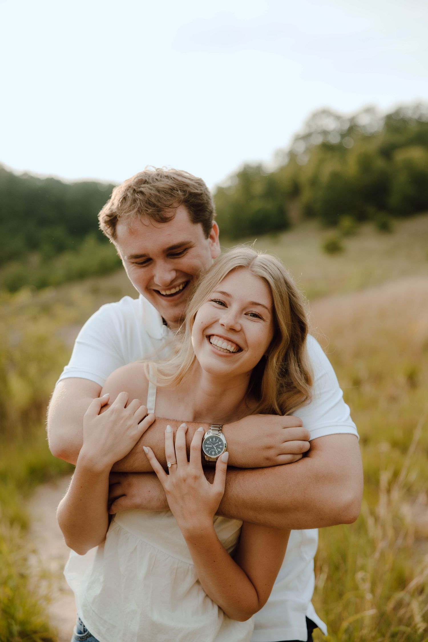 A joyful couple embraces in a sunny meadow, the man hugging the woman from behind as both laugh happily.
