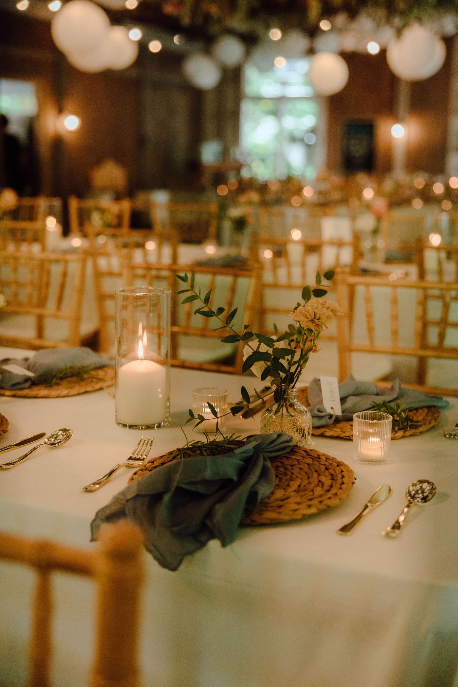 Elegant wedding reception table with gold chiavari chairs, candlelight, woven chargers, dusty blue napkins, and greenery centerpieces.